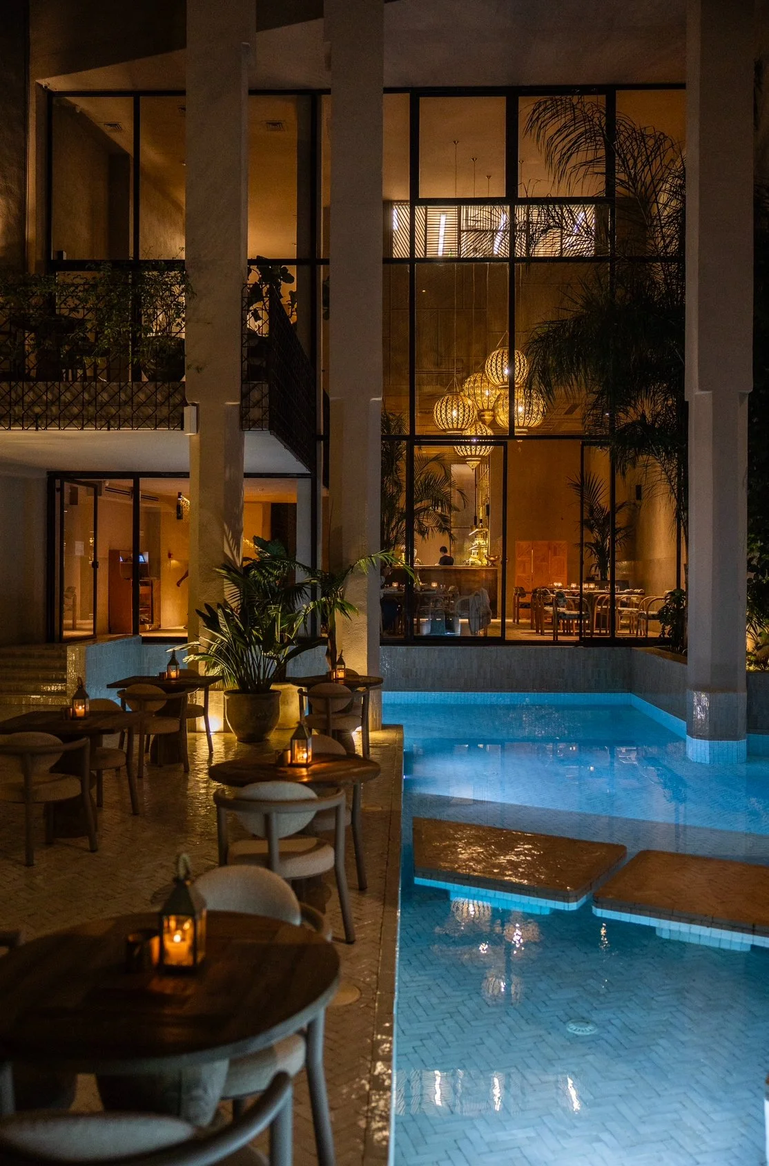 Naama restaurant Marrakech. Nighttime view of a luxurious indoor pool area with tables and chairs, dimly lit by lanterns on the tables. The background features large windows revealing an elegant interior with decorative lights and plants.