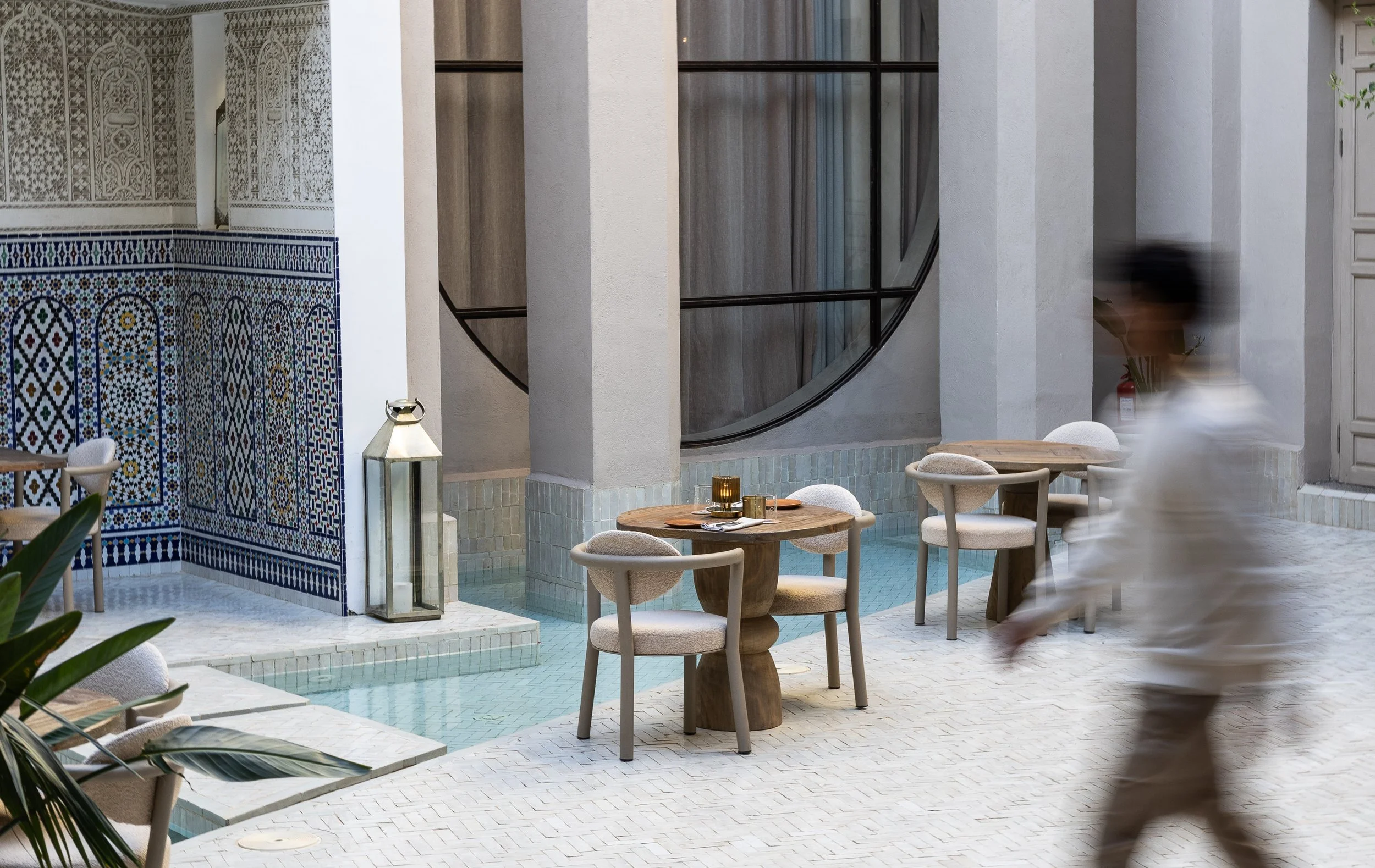 Naama restaurant Elegant outdoor patio with tiled walls, wooden tables, chairs, and small pool. Decorative lanterns and mosaic details enhance the setting. A person is walking past, slightly blurred.