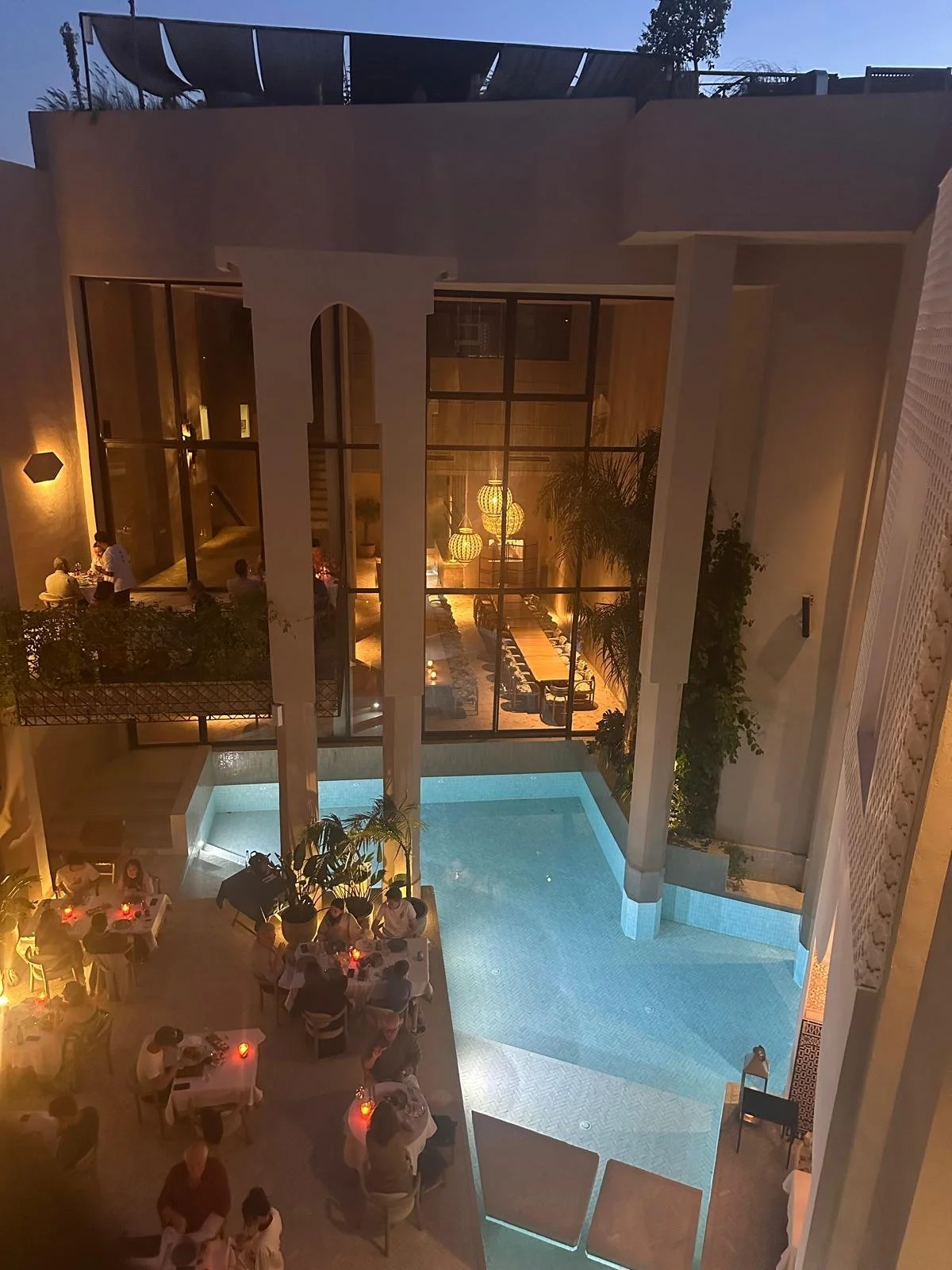 Naama restaurant Marrakech. A nighttime view of a luxurious dining area beside a swimming pool, with tables set for dinner. The setting features elegant architecture with large glass windows, decorative lighting, and lush greenery.