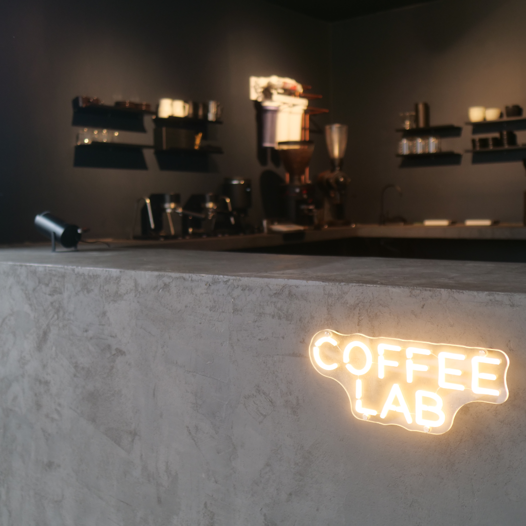 The image shows a modern coffee shop counter with a neon sign that reads 'COFFEE LAB' on a textured concrete surface, and dark walls with shelves holding coffee cups and equipment in the background.