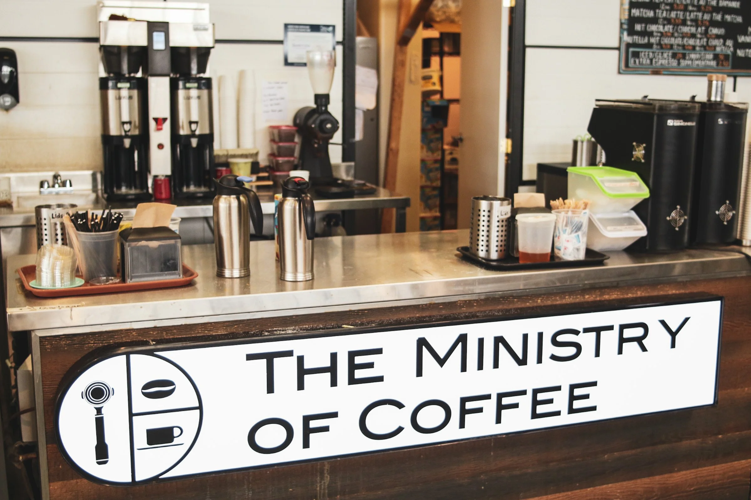 Counter at a coffee shop called The Ministry of Coffee with coffee brewing equipment, cups, and supplies.