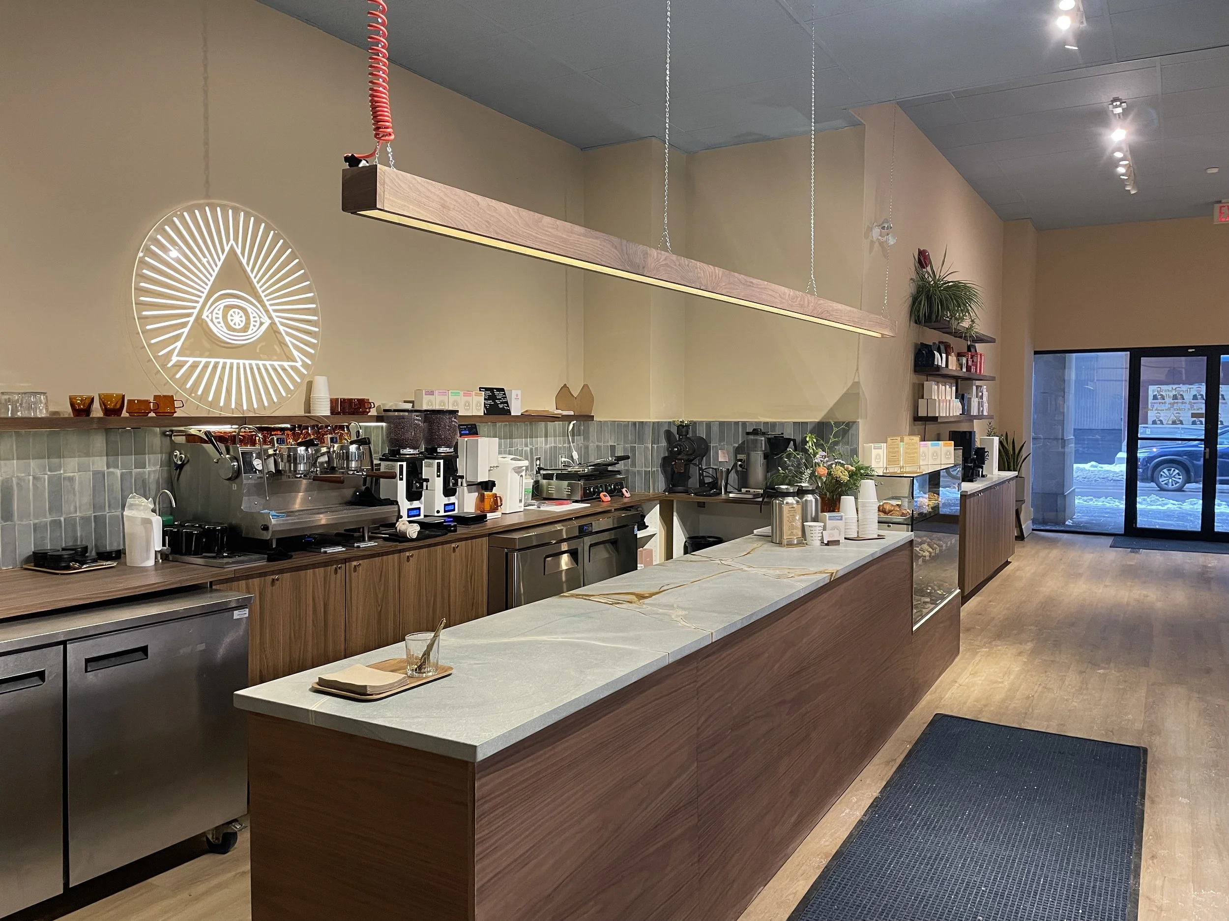 Coffee shop interior with a counter, coffee machines, a neon sign of an eye within a triangle with rays, and a sliding glass door at the back showing a snowy outdoor scene.
