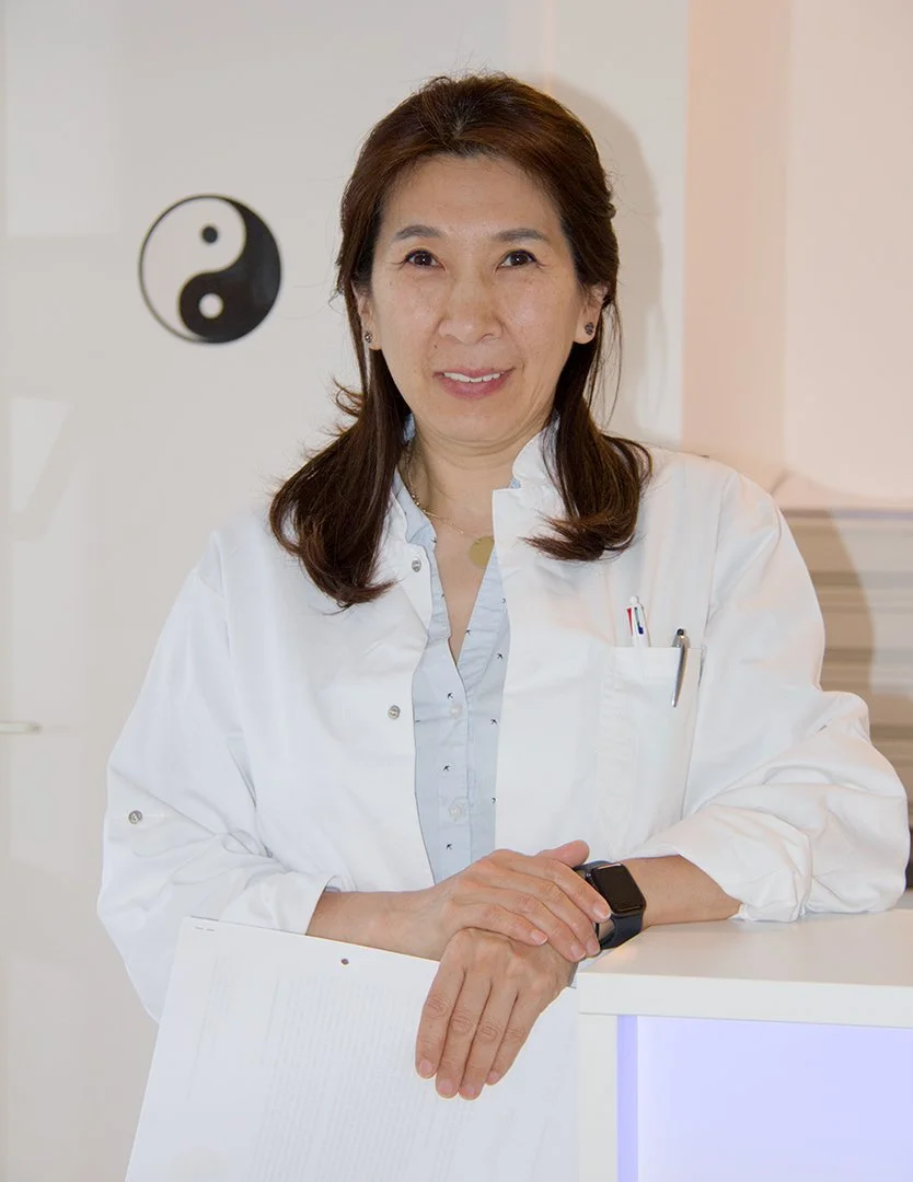 Ärztin mit weißen Kittel, steht in Praxis mit Yin-Yang Symbol im Hintergrund.