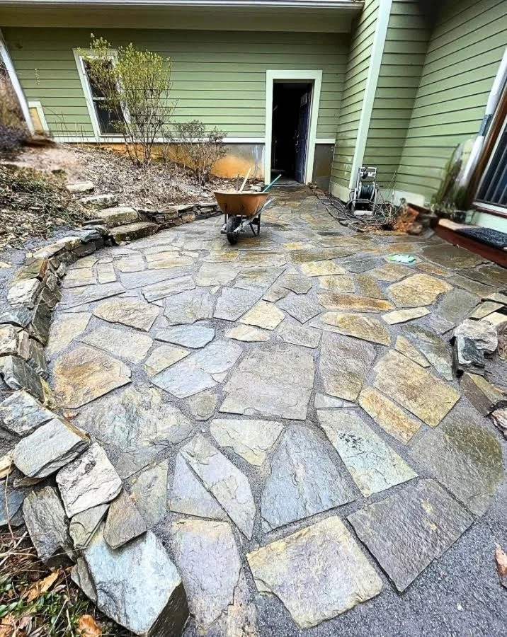 A natural stone patio build with irregularly shaped stones in various hues of grey and some yellow. The patio is bordered by natural stone retaining wall as well. All attached to a green house in Swannanoa, NC