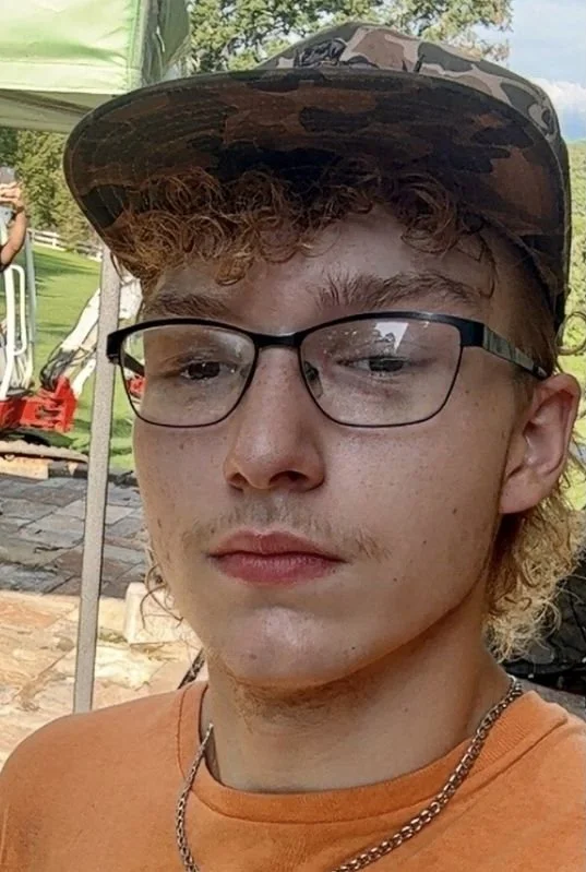 A young man with curly reddish hair wearing black-rimmed glasses, a camouflage cap, and an orange shirt taking a selfie outdoors. In the background, there are trees, a tent, and some outdoor equipment.