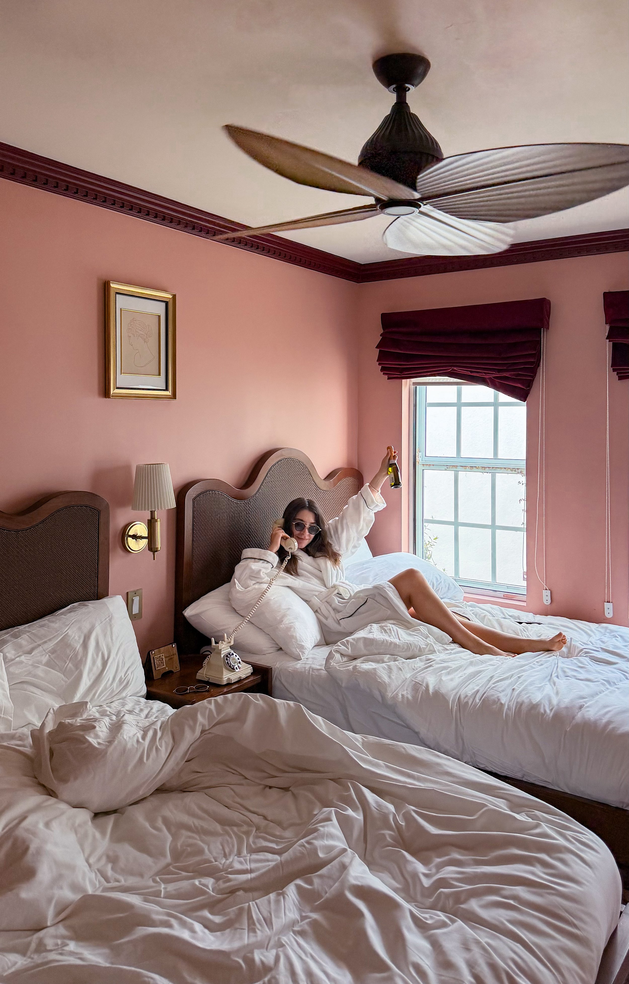 A woman sitting on a bed in a pink bedroom, talking on the phone, wearing sunglasses, holding a beer, and raising her arm. Two beds with rumpled white sheets, a vintage rotary phone, and a framed line drawing on the wall are visible. The room has a window with red drapes and a ceiling fan.