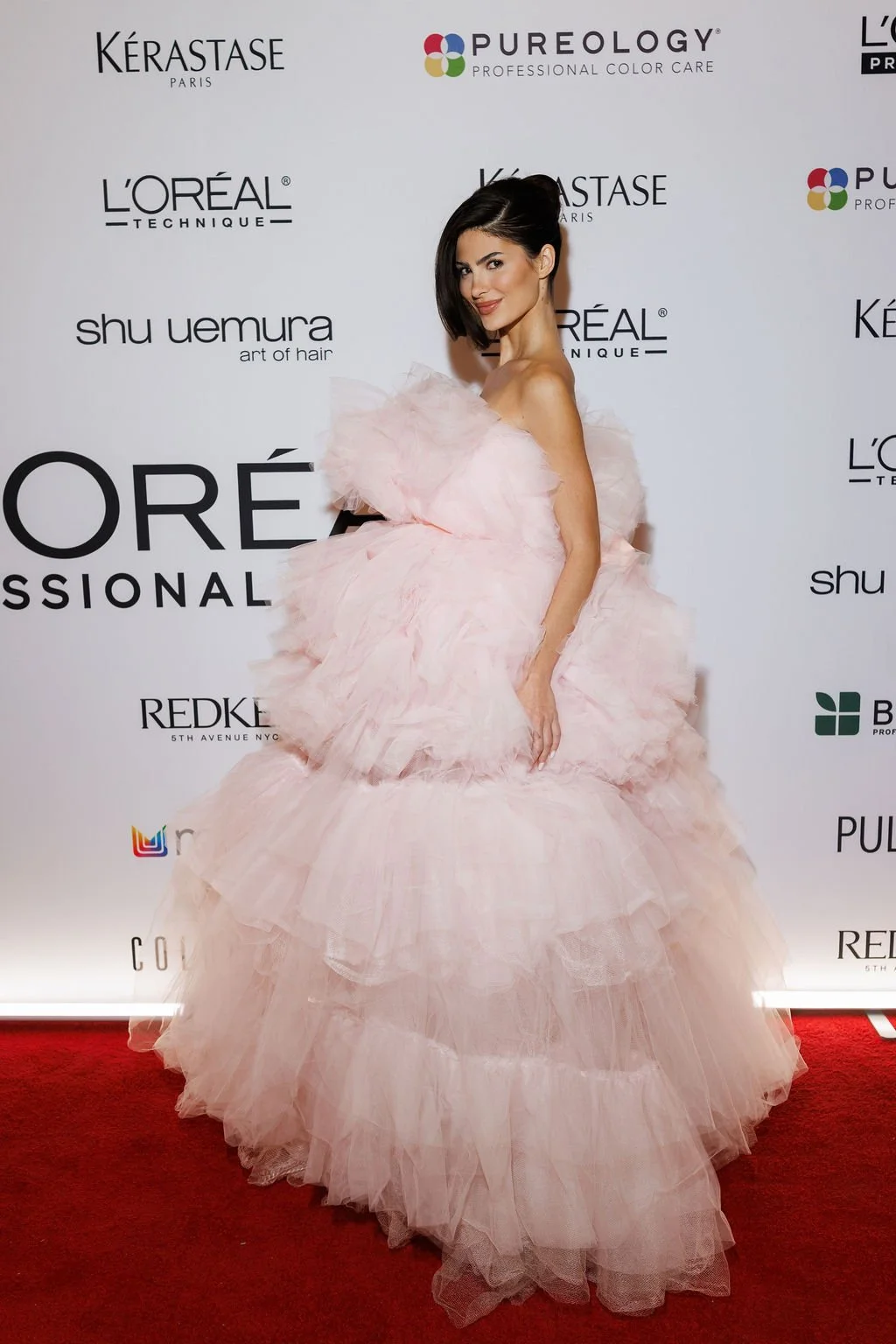 Woman in a pink, ruffled, strapless gown standing on a red carpet with a white backdrop featuring logos of beauty and haircare brands.