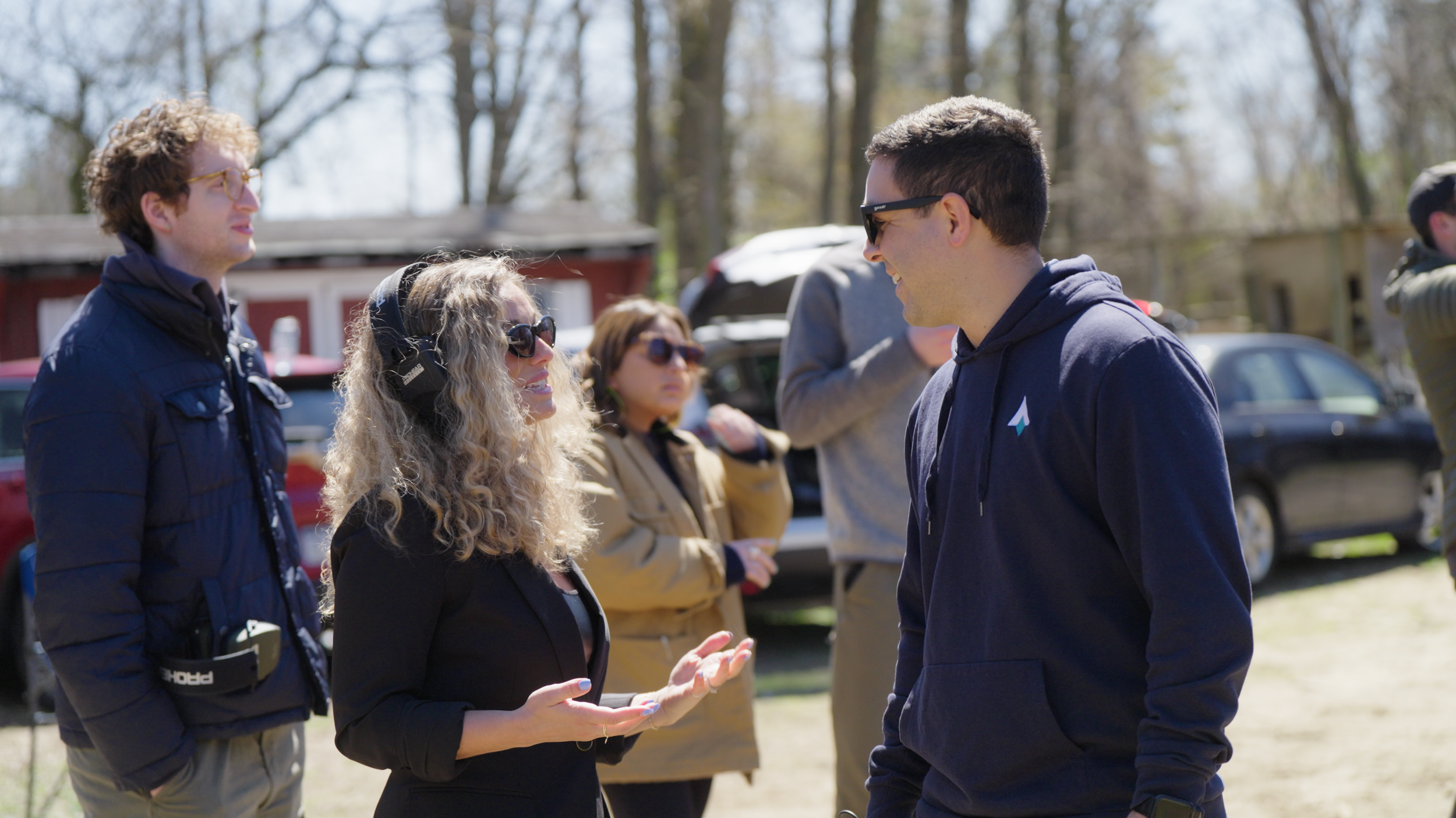 A group of people outdoors engaging in conversation, with trees and parked cars in the background.