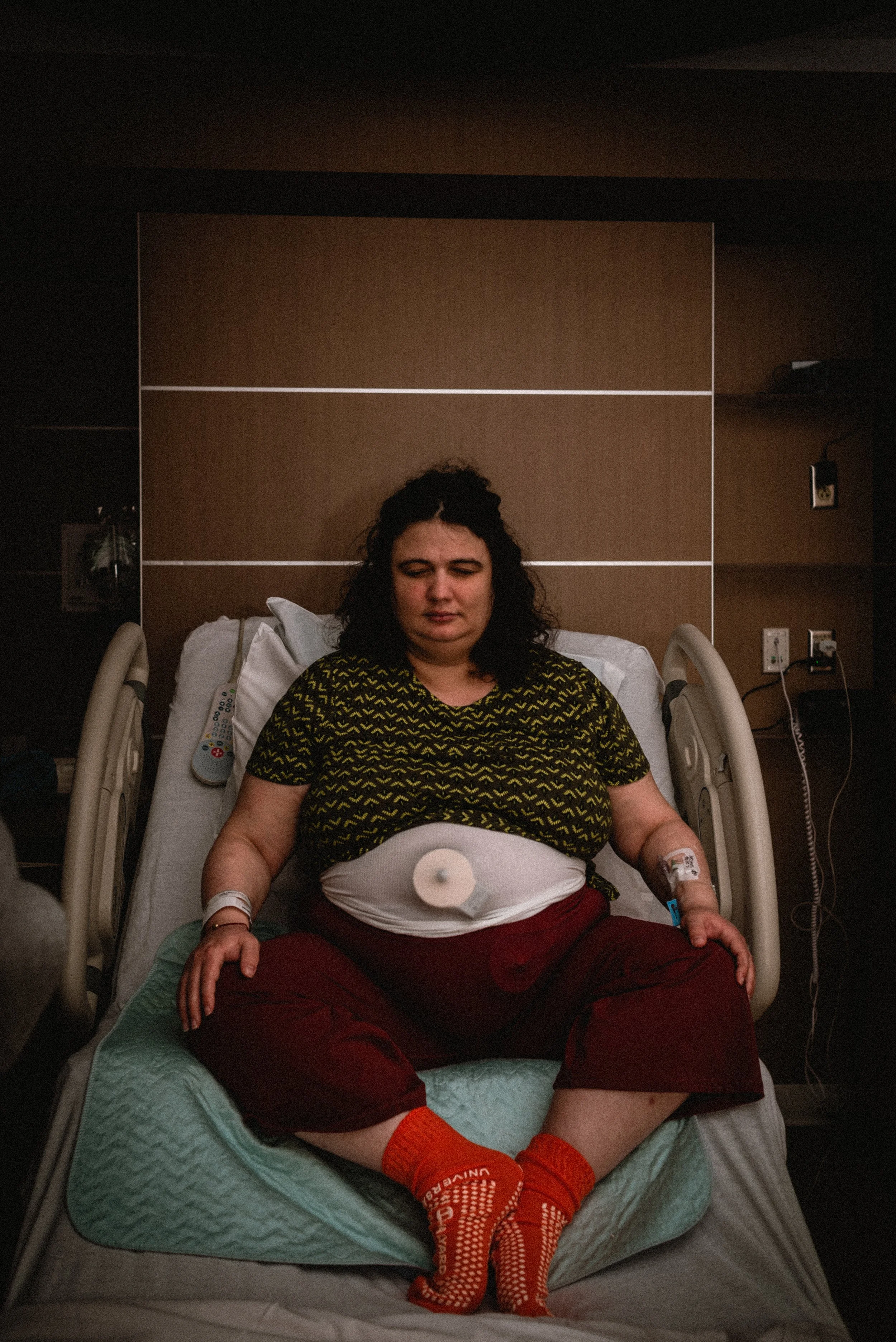 Lucy sits on a hospital bed with a peaceful expression on her face. Her hands rest gently on her knees, sitting in butterfly pose, while her feet touch together. Fetal monitors are visible through the white mesh band on her belly, holding them in pl