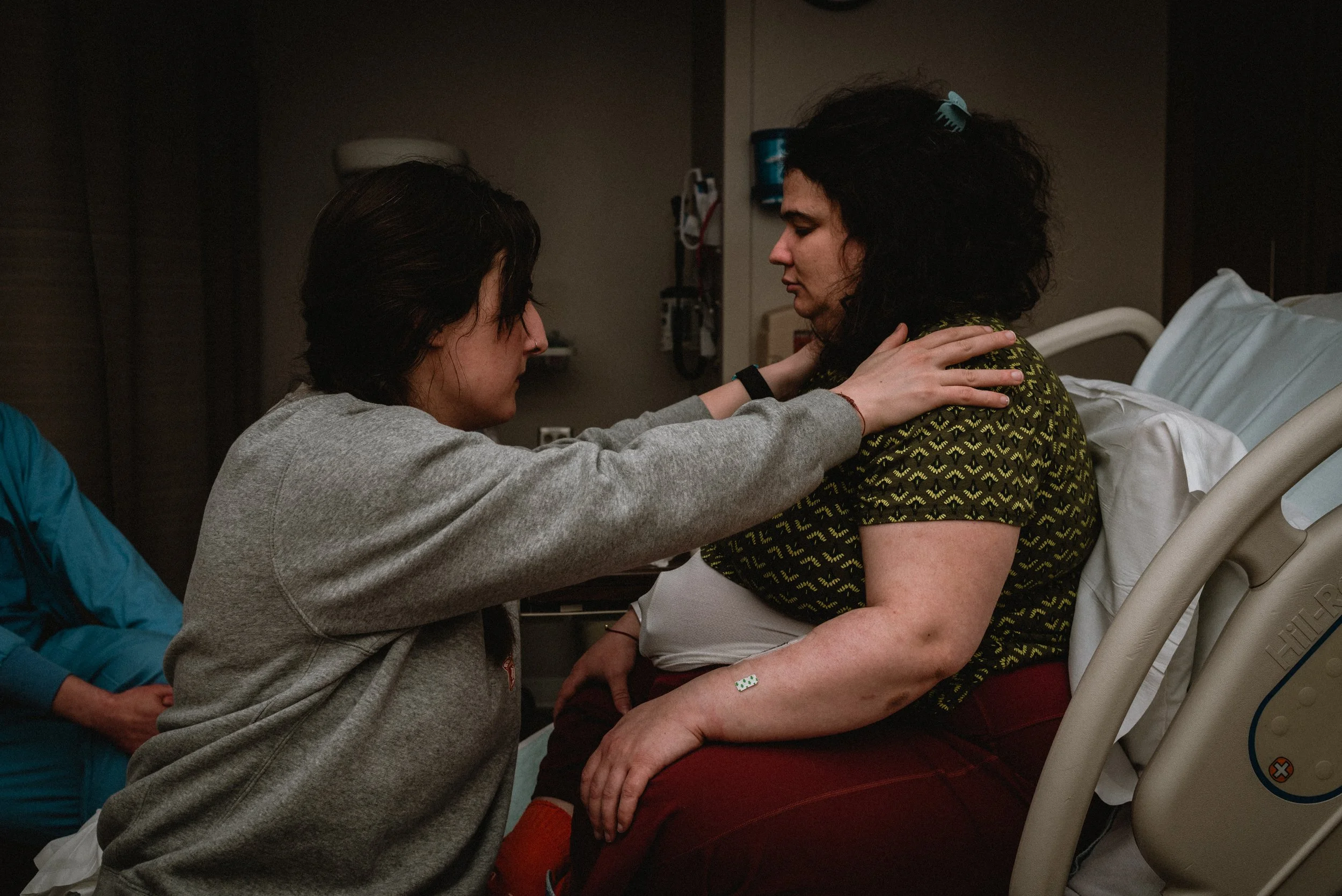  Lucy is seen from the side, hands on her knees, while she sits in a hospital bed. Ale sits sideways on the bottom of the bed, which is lower than Lucy. Ale’s hands are gently pressing Lucy’s shoulders away from her ears, while she looks intently at 