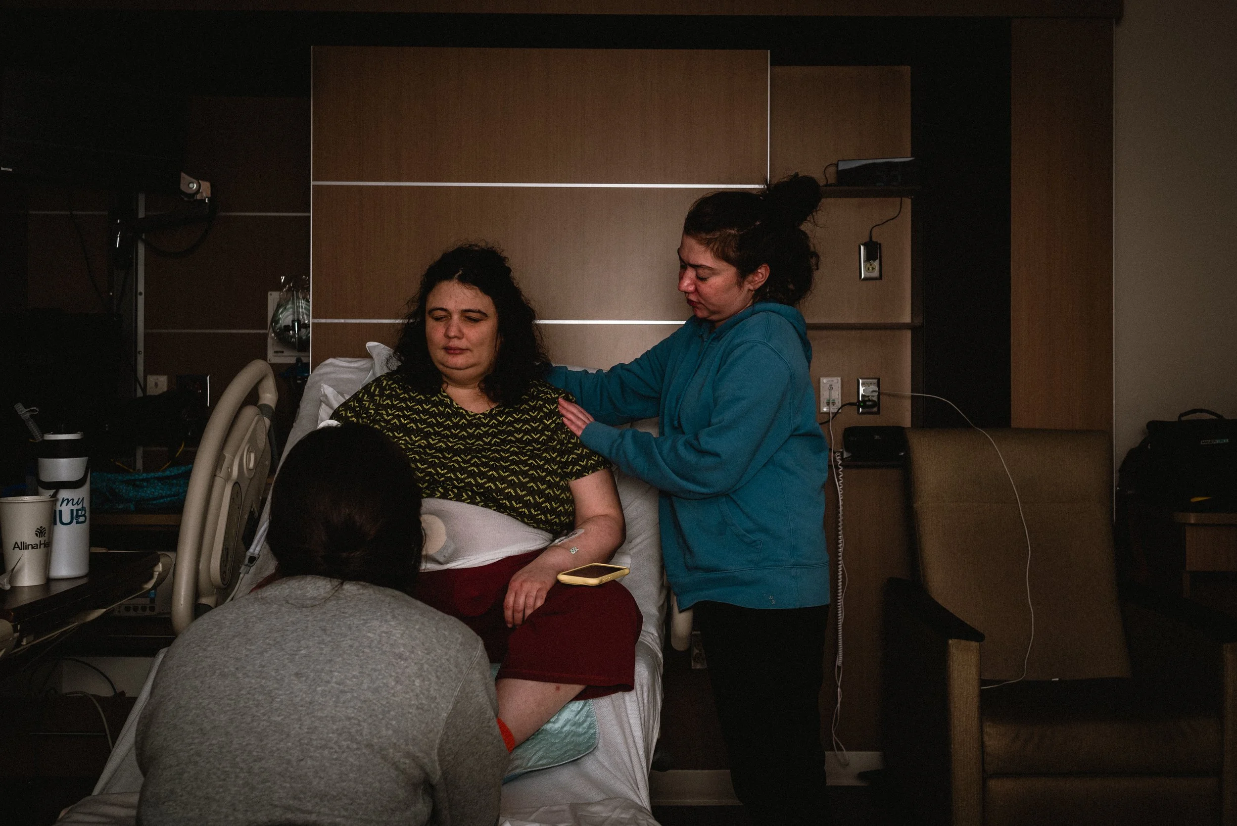  Lucy sits in a hospital bed with her phone resting on her knee. Her sister Rose is standing to her left, arms draping around Lucy to keep her relaxed during a contraction. Ale’s back is to the camera as she sits on the bottom of the bed, holding Luc