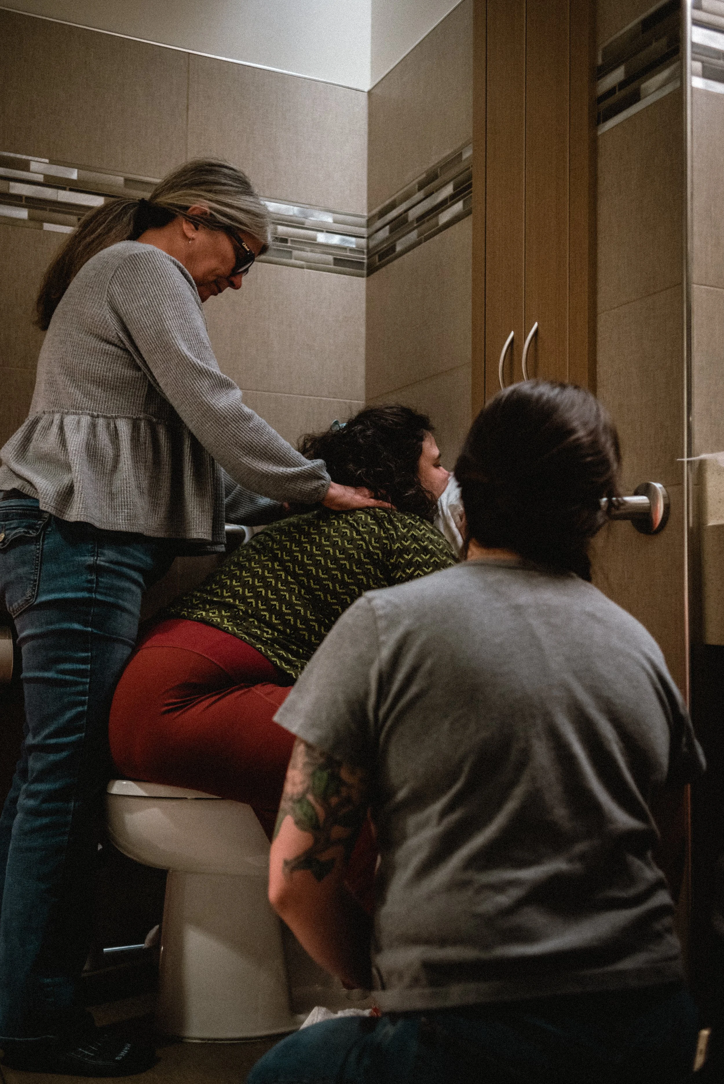  Lucy sits backwards on the toilet, resting her face into a pillow that sits on the back of the tank. Meg is standing behind her, staring down and her while she rubs her shoulders. Ale’s back is to the camera as she kneels in front of Lucy, looking p