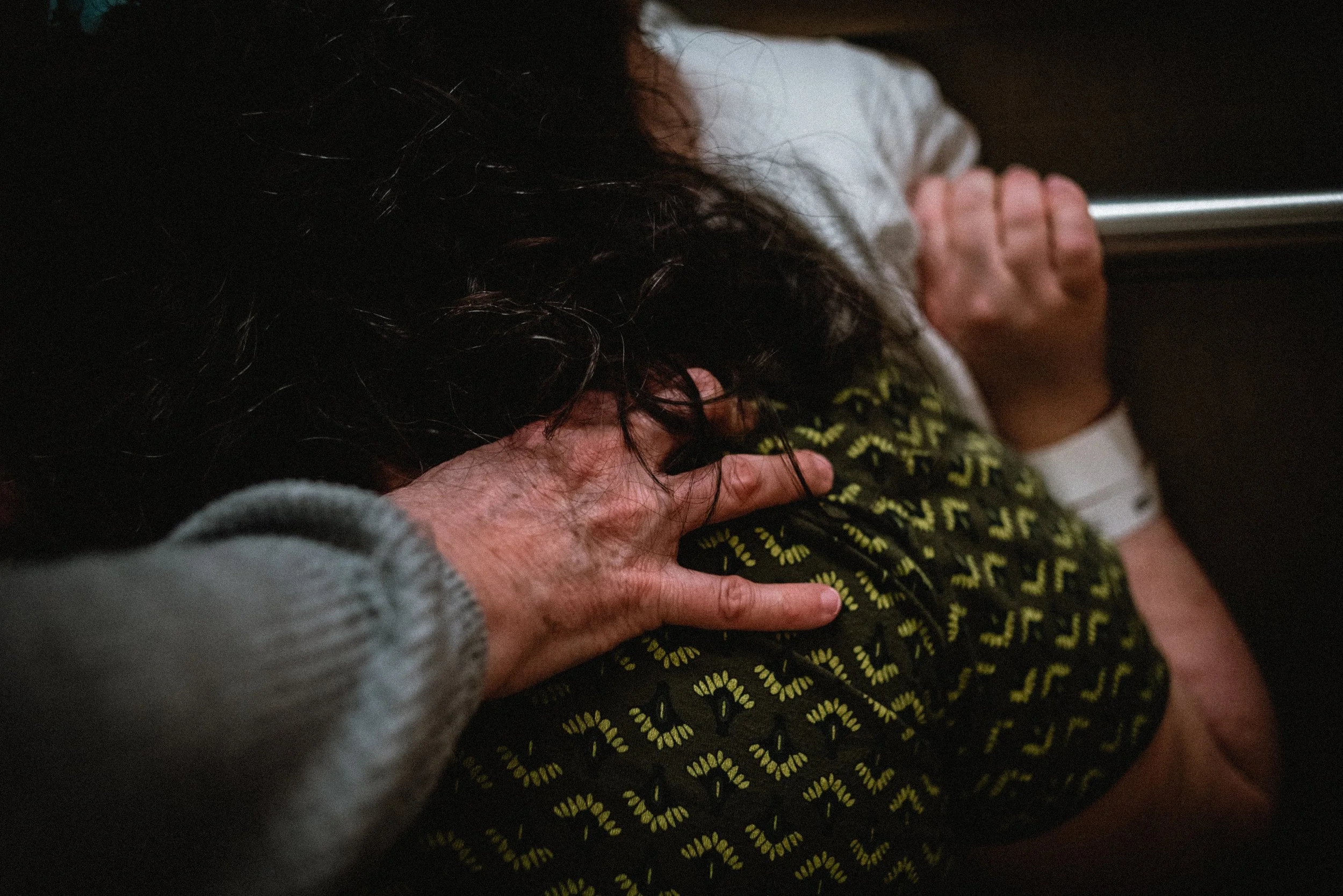  A close up image of Meg’s gentle and grounding hand. Her fingers are flexed on Lucy’s shoulder. Lucy’s dark curls fall down her shoulders, gently settling on Meg’s fingers. Hazy in the background, Lucy’s hand can be seen gripping a metal hand rail. 