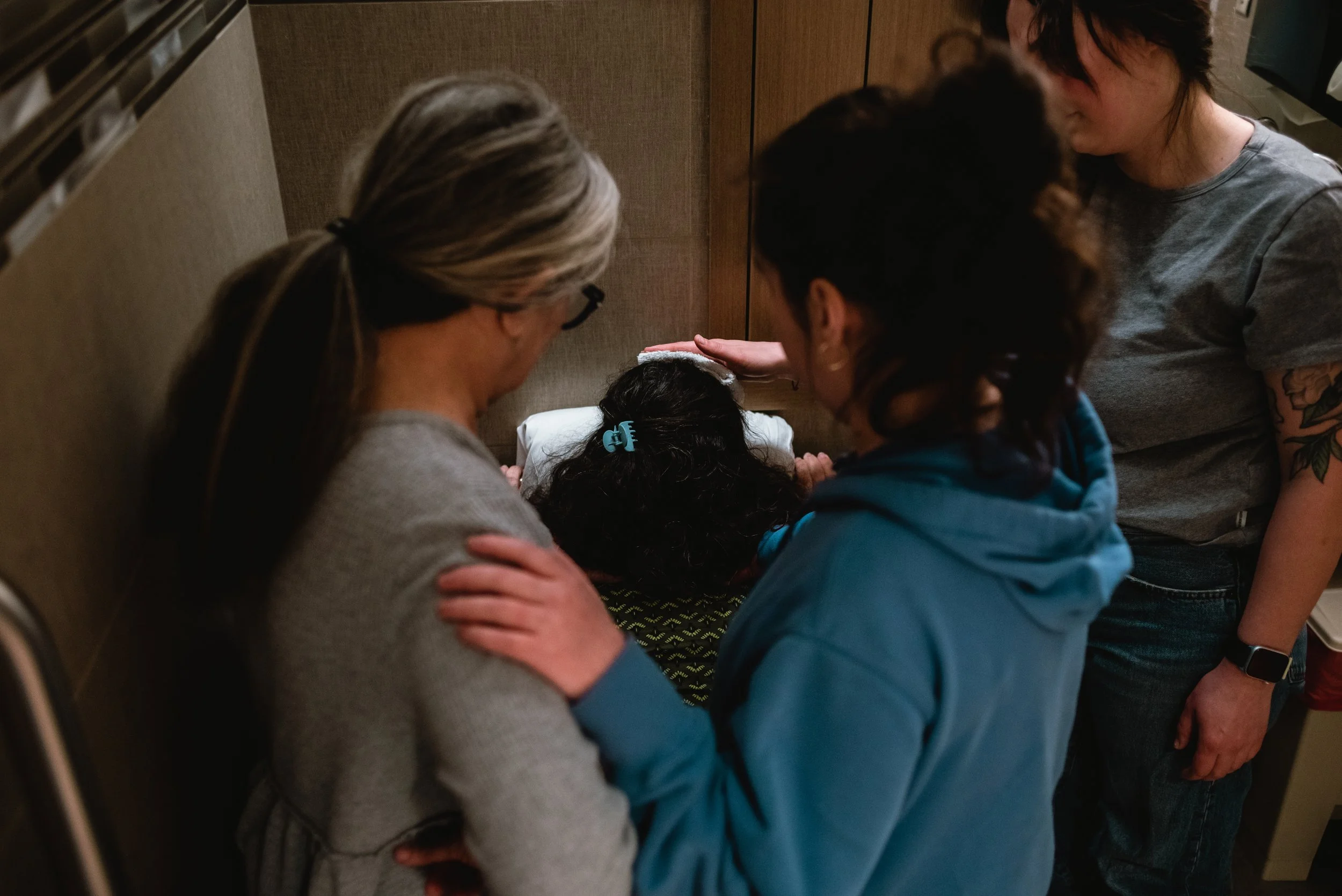  Meg and Rose are in the center of the photo, Rose’s hand resting on Meg’s shoulder. They both look down out Lucy, who is framed by their bodies. Lucy rests backwards on the toilet. Ale stands off to the right, with her arm outstretched to hold a coo