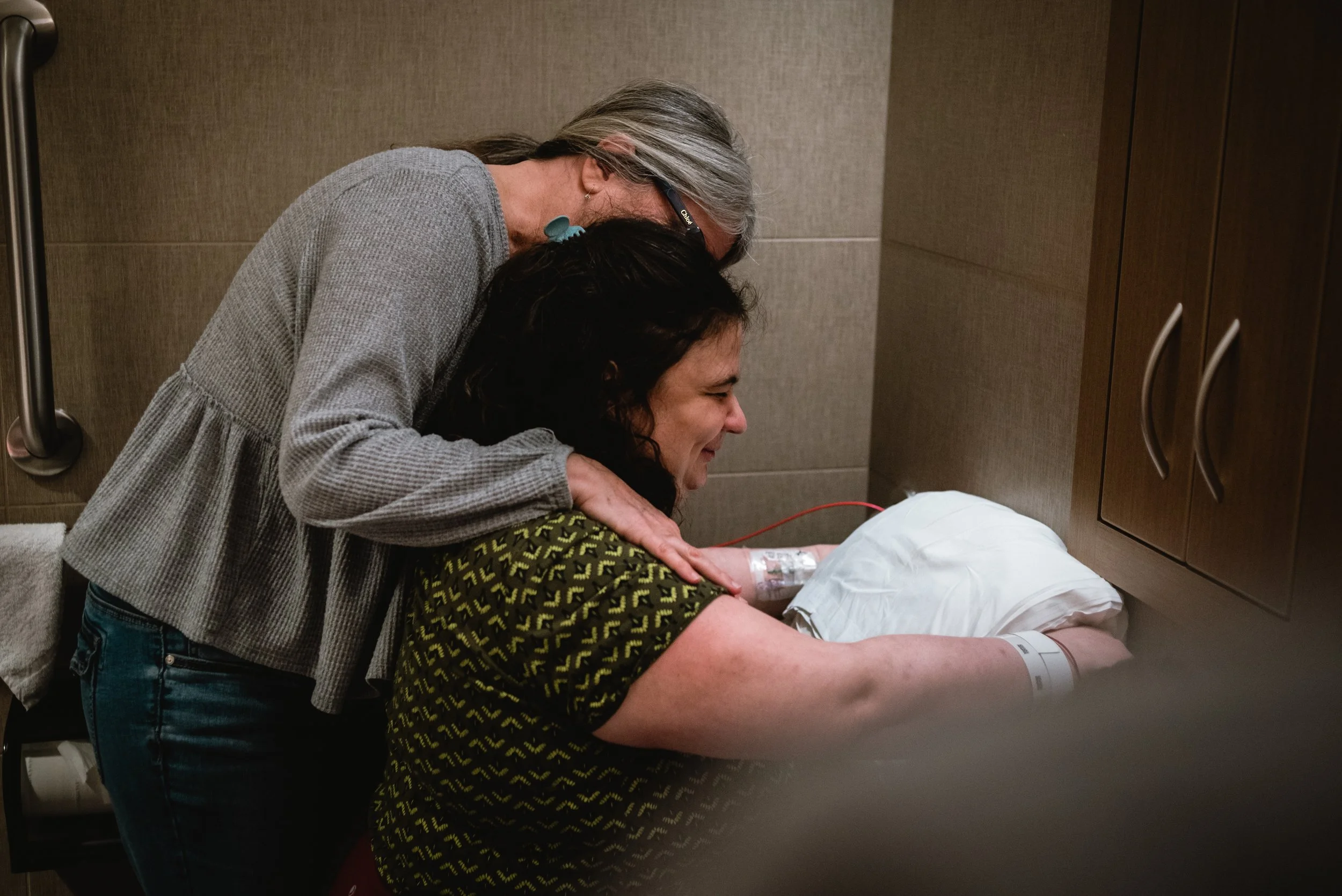  Meg and Lucy are seen waist-up from the side, as Meg leans forward over Lucy to hug her, her face obscured by Lucy, who is smiling. Lucy is facing backward on the toilet, her hands gripping the handrail on either side of a pillow, which rests on the