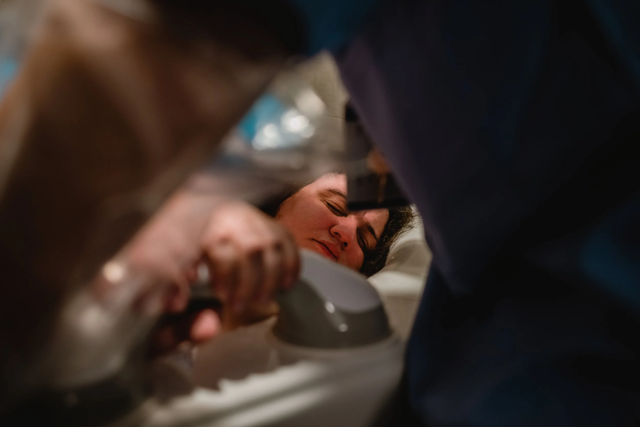  Lucy is in the center of the photo, her eyes closed as she rests her head on a pillow lying on the edge of the bathtub. Her hand grips a handrail attached to the side of the tub. Lucy’s head and hand are framed by the outline of a nurse’s side and a