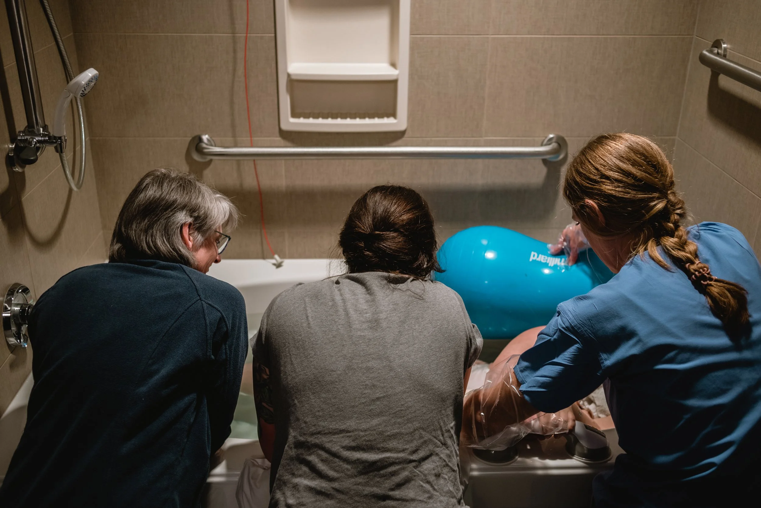  Janine, Ale, and a nurse rest their elbows on the edge of a bathtub, as the look toward Lucy, their backs to the camera. A large blue peanut-shaped birth ball can be seen, supporting Lucy’s position in the tub. While Lucy isn’t visible in the image,