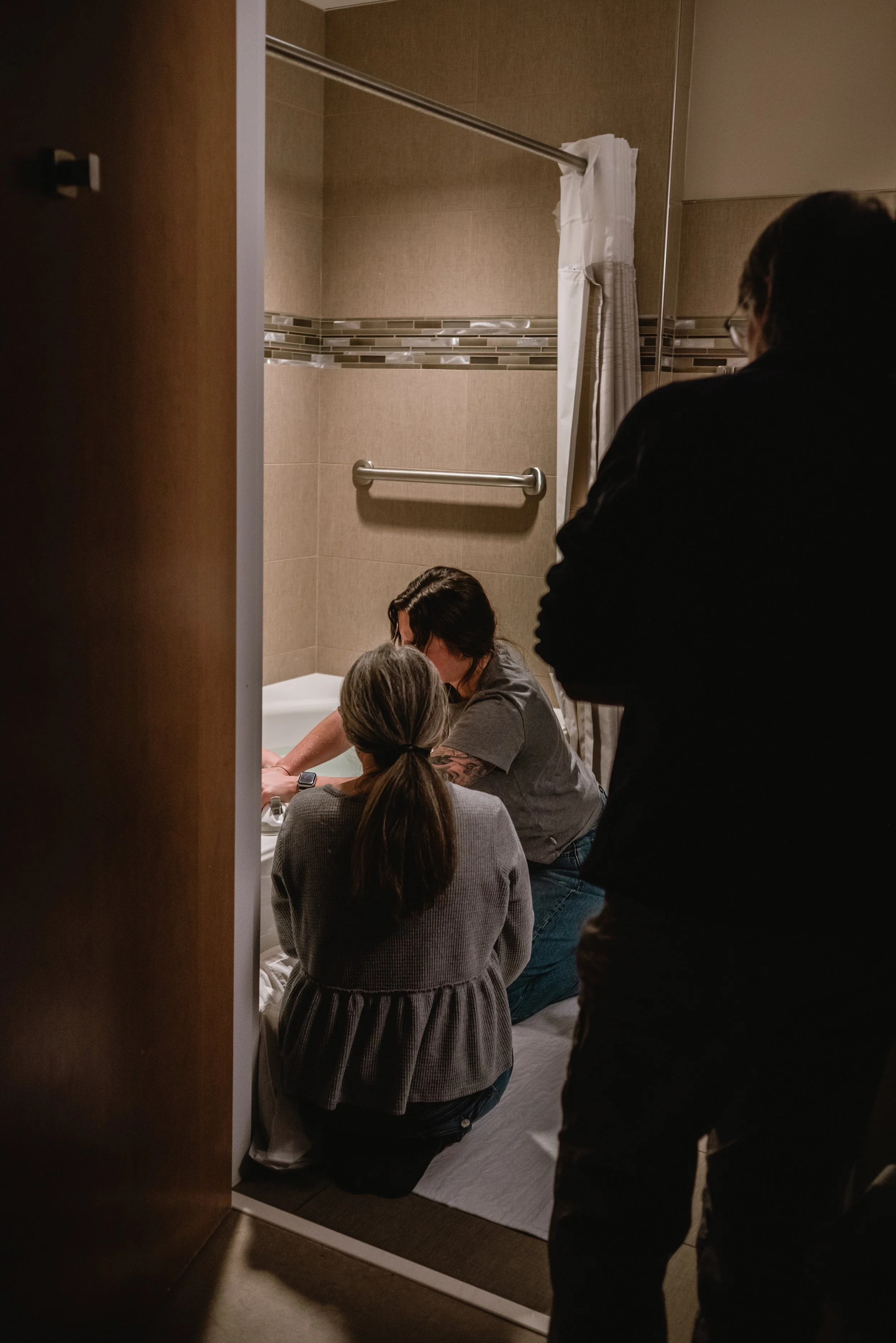  Janine’s back is to the camera as she leans against the bathroom door frame, peering in toward Meg, Ale, and Lucy. Meg is kneeling on her heals, facing the tub, while Ale leans over the tub to make adjustments for Lucy’s comfort. Lucy is hidden behi