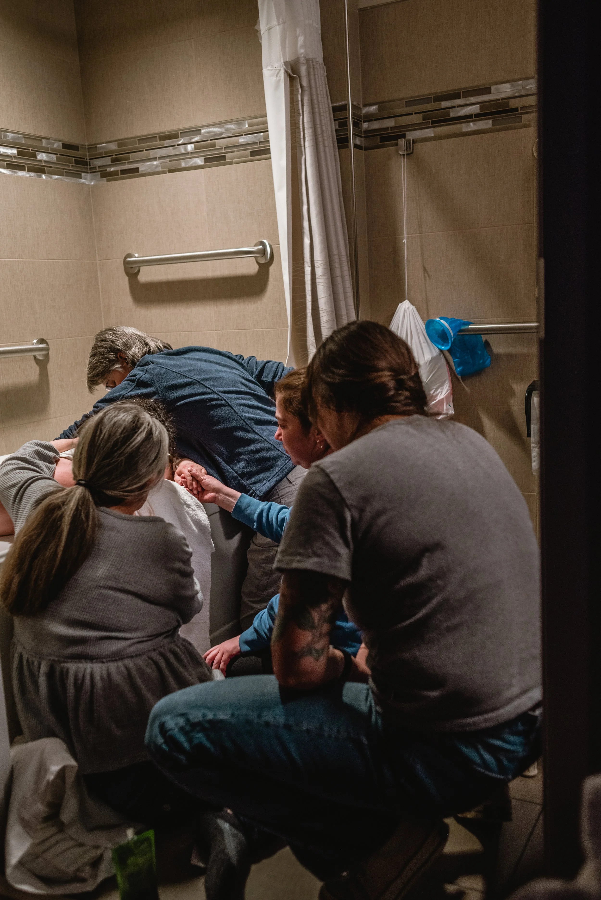  There are many busy bodies attending to Lucy, who sits in the tub and is shielded by their work. Meg, with her back to the camera, kneels while reaching to Lucy. Janine stands, bent over the tub to press on Lucy’s back during a contraction. Ale squa