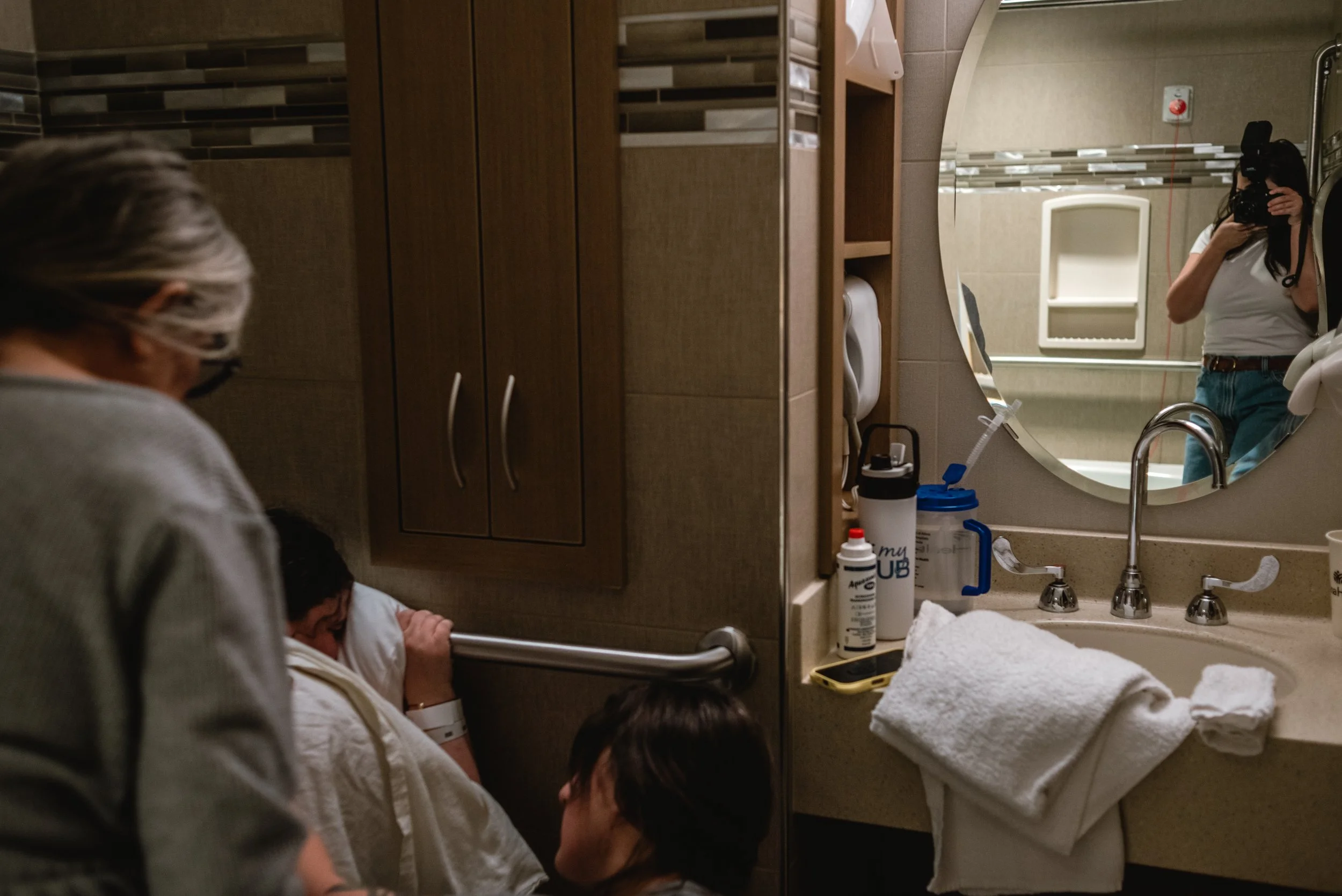  Gina is reflected taking a picture in the bathroom mirror, off to the right edge of the photograph. In the left edge of the image, Meg can be seen rubbing Lucy’s back as she sits backwards on the toilet, draped in a warm blanket, and gripping the ha