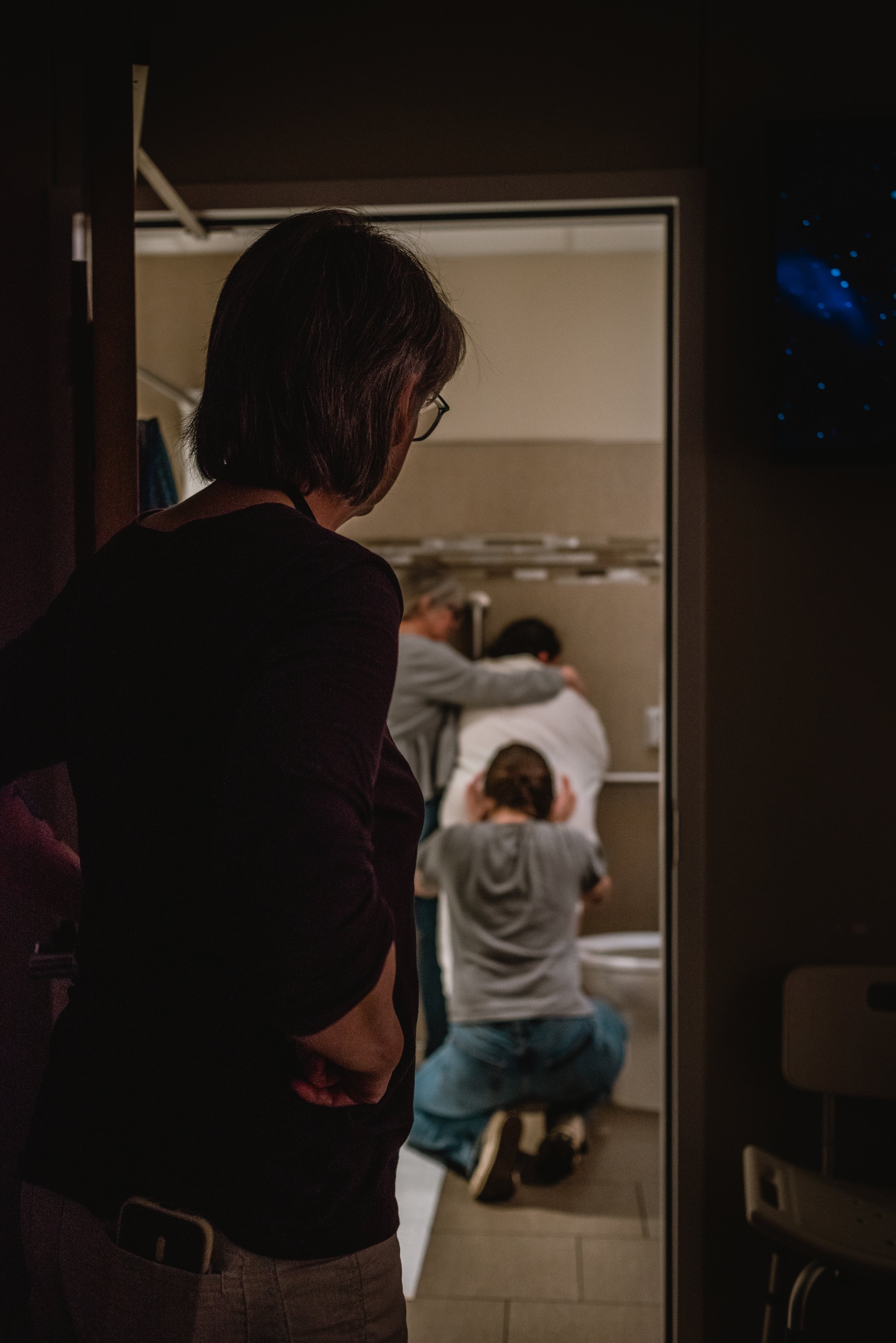  Janine stands outside the bathroom, the dark birth room casting her in shadow on the right side of the image. She has her hand relaxed at her hip as she looks into the bathroom. Lucy stands framed by the doorway, facing the wall during a contraction