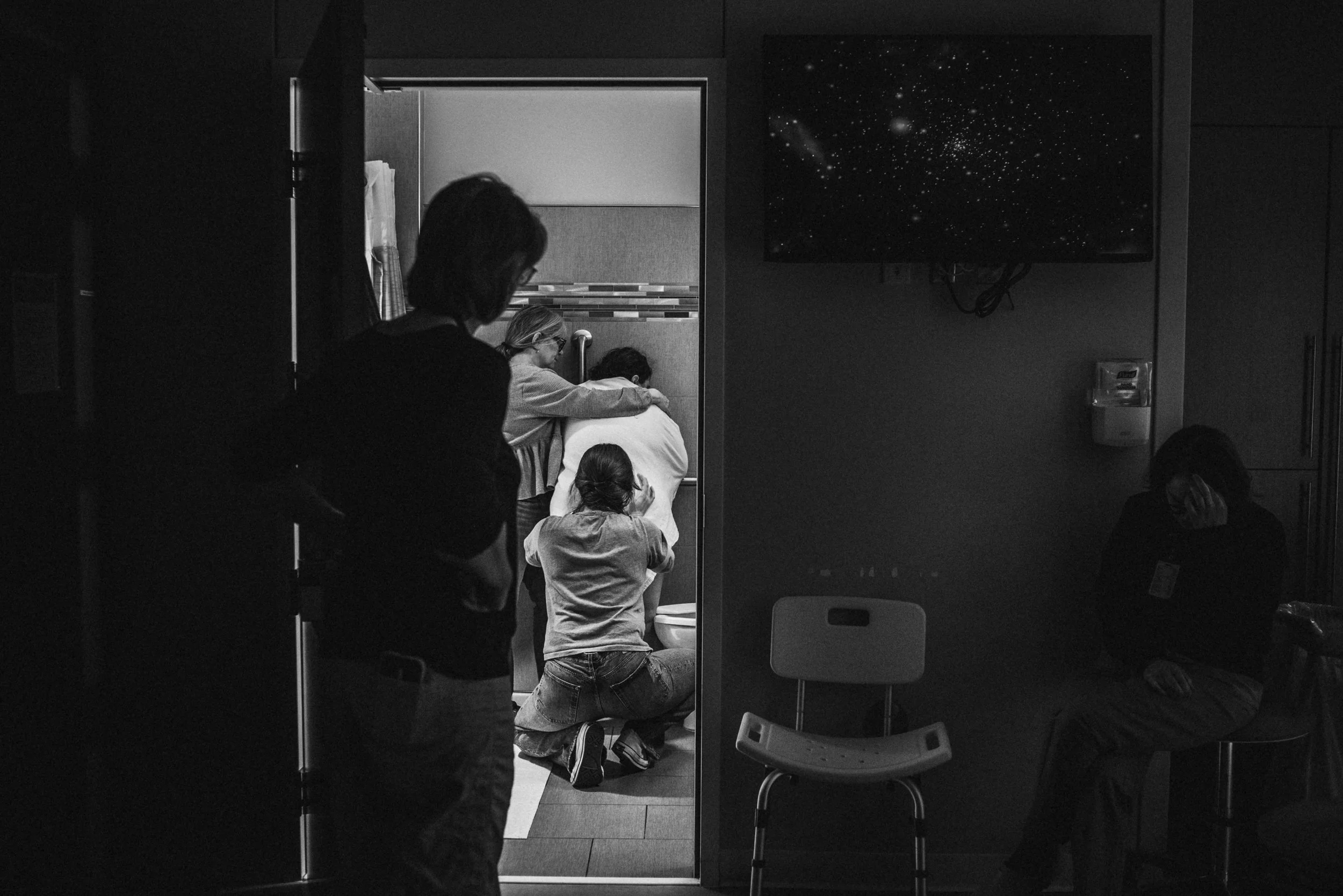  A black and white image shows Janine peering into the doorway from the birth room. Meg stands by Lucy, who is facing the wall, with her arms on Lucy’s shoulders. Ale kneels behind Lucy, squeezing her hips. To the left of the door, nurse midwife Kati