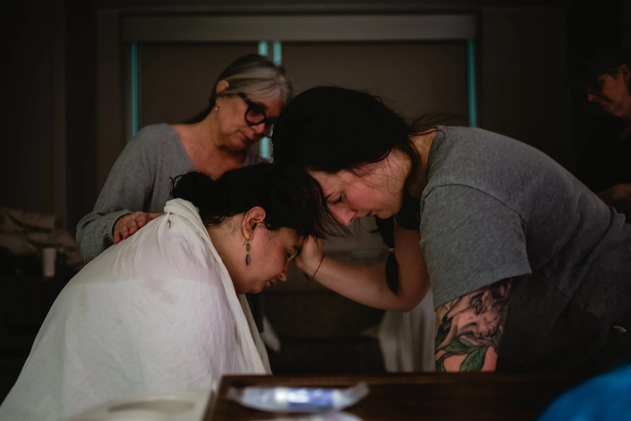  Lucy is draped in a blanket and sits on a hospital bed, her head bowed. Ale stands in front of her, her right arm on her shoulder, her head bent and forehead touched to Lucy’s. Meg stands to Lucy’s left, rubbing her back and focusing on her daughter