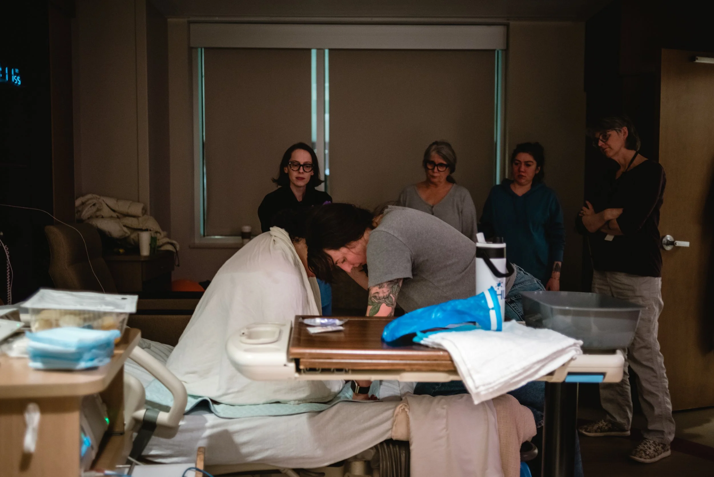  A wide image of Lucy, draped in a white blanket, sitting on a hospital bed, her head bowed forward. Ale kneels in bed with Lucy, her right arm around her and head on Lucy’s shoulder as she whispers to her. Behind them, nurse midwife Katie, Meg, Rose