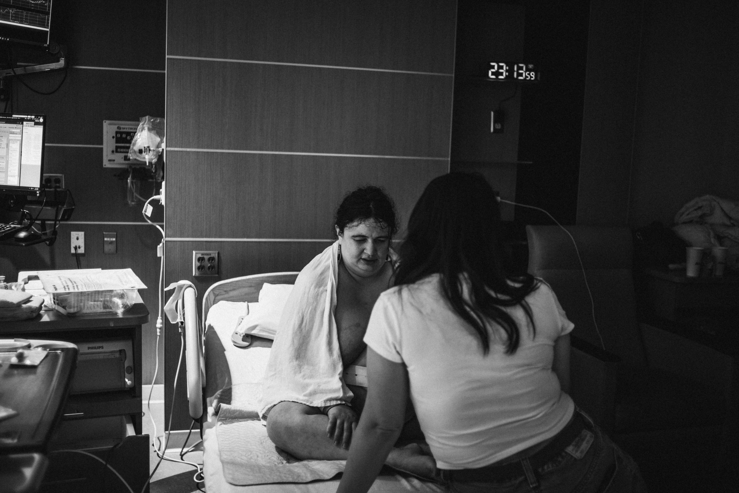  A black and white image of Lucy sitting in a hospital bed, draped in a white blanket and facing the camera, her legs in a criss/cross position. Gina leans on the bed in front of her, her back to the camera as she speaks with her. Light illuminates t