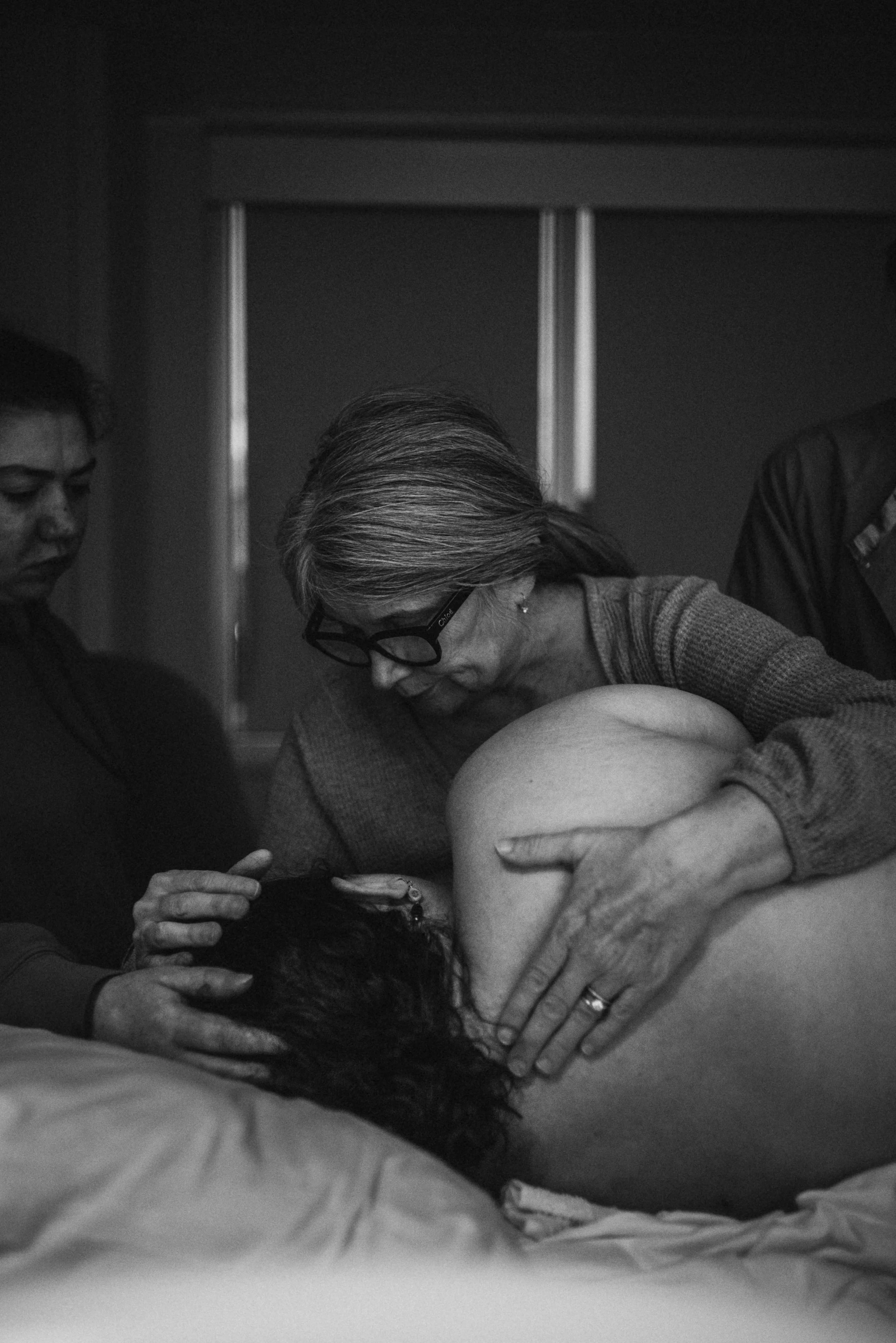  A black and white image of Meg curled over Lucy, who is on her side with her back to the camera. Meg’s hand is on Lucy’s back, while her other hand cards through Lucy’s curls, meeting the touch of Rose’s hand, who stands above Lucy to the left.  