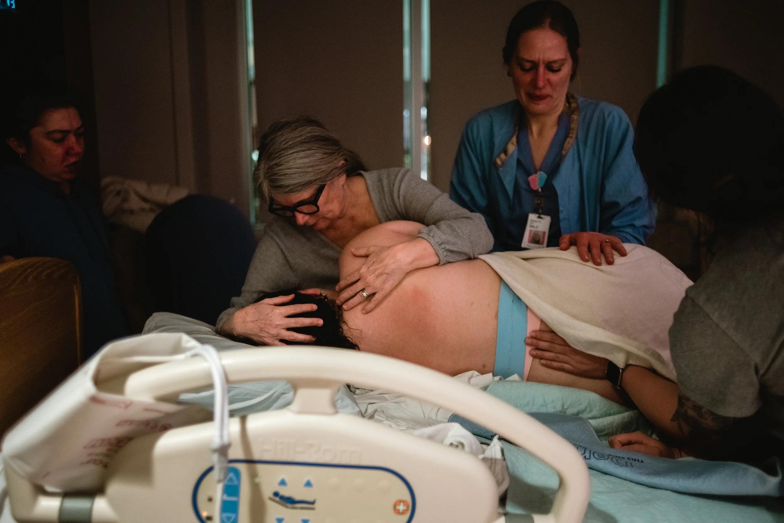  Meg holds her daughter with both arms as she stands over her. Lucy lays in bed with her back to the camera, while Ale leans in from the right side of the image to place her palms on Lucy’s lower back. Rose stands over, watching Lucy from the left. N