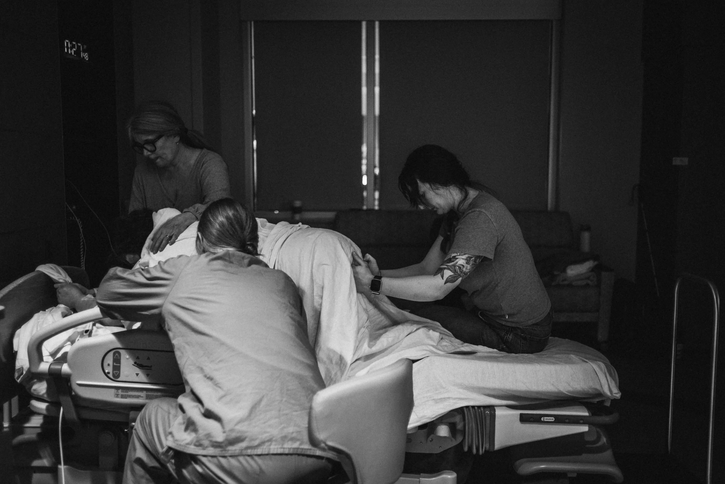  A black and white image focuses on Ale, sitting sideways on the bottom of a hospital bed, using both fists to press into Lucy’s sits bones. Lucy is draped in a large white blanket, on hands and knees and she copes through a contraction. Nurse Caryn’