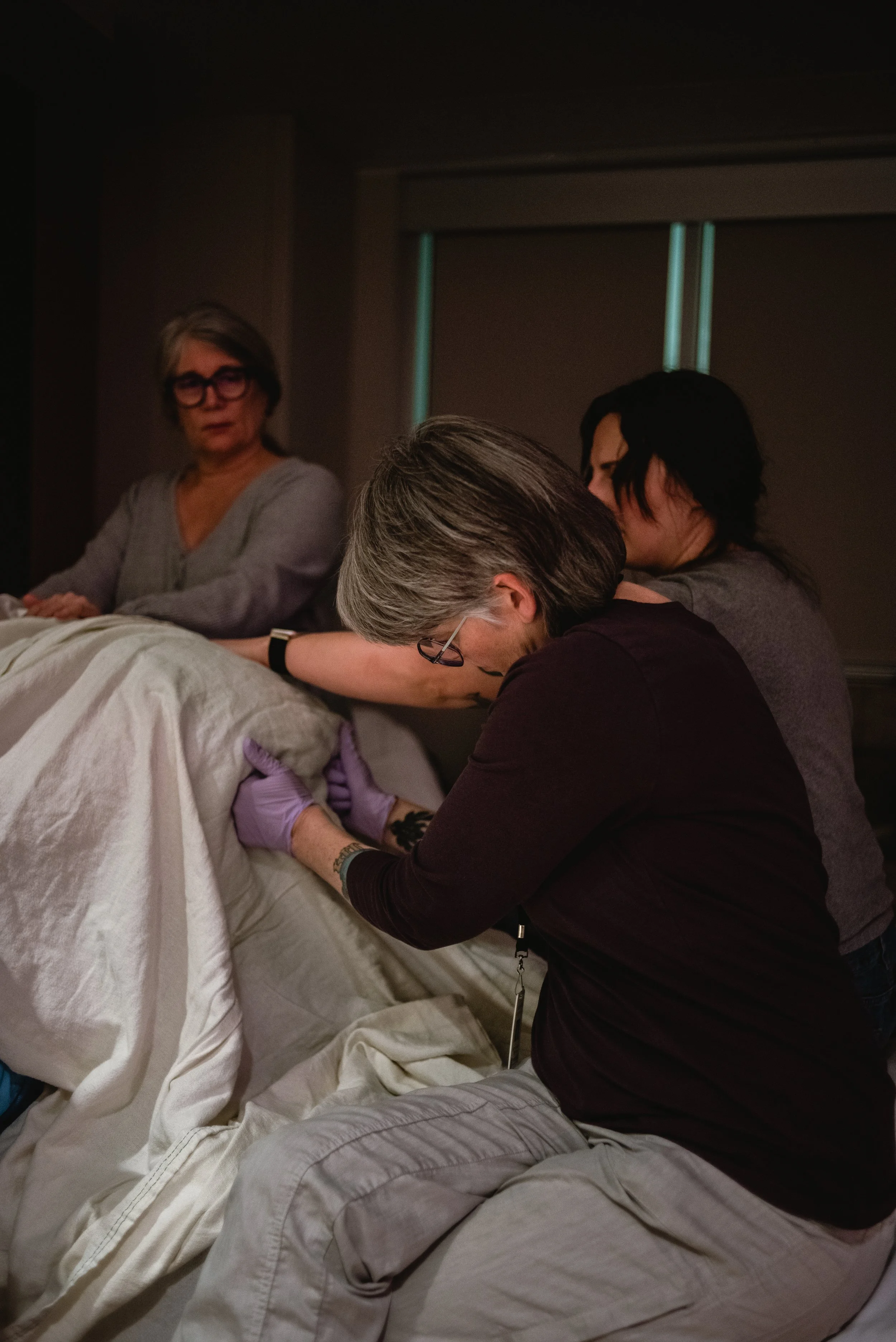  Janine is the the focus, at the center of the image. Her hands are covered in purple nitrate gloves as she presses against Lucy’s sits bones. Her head is bowed, focused on the work at hand. Ale rests on the end of the bed behind Janine, her arm rest