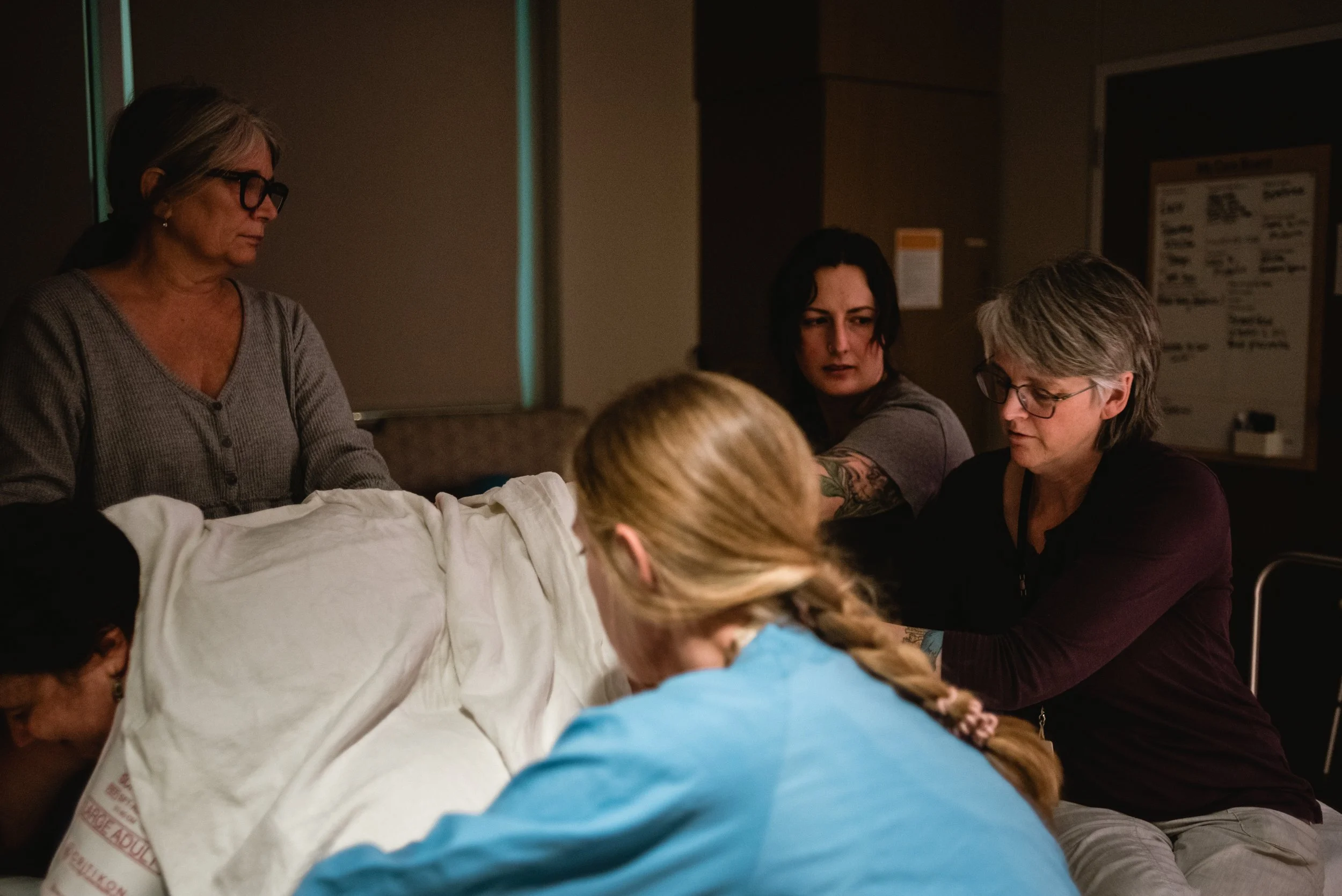  The image is taken from the head of Lucy’s bed, where she rests on her hands and knees during a contraction. Her body is covered in a white blanket. Janine is at the bottom of the hospital bed, pressing on Lucy’s sits bones, while Ale sits beside he