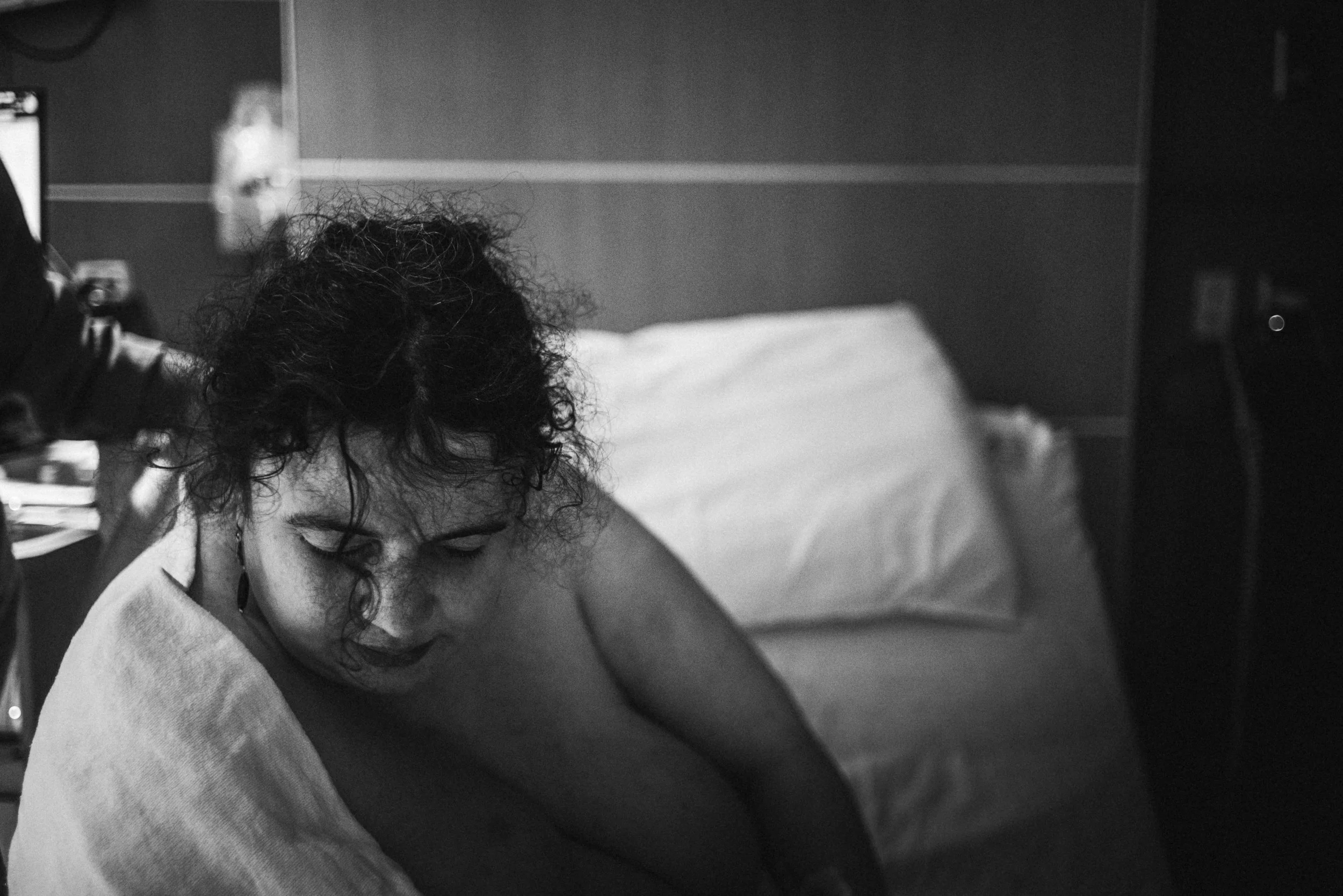  A black and white image of Lucy from the chest up, in between contractions. A white blanket slips from her shoulders. Her head is cast down, eyes closed and resting. The hard work is clear through her mussed curls. A single ringlet falls in front of