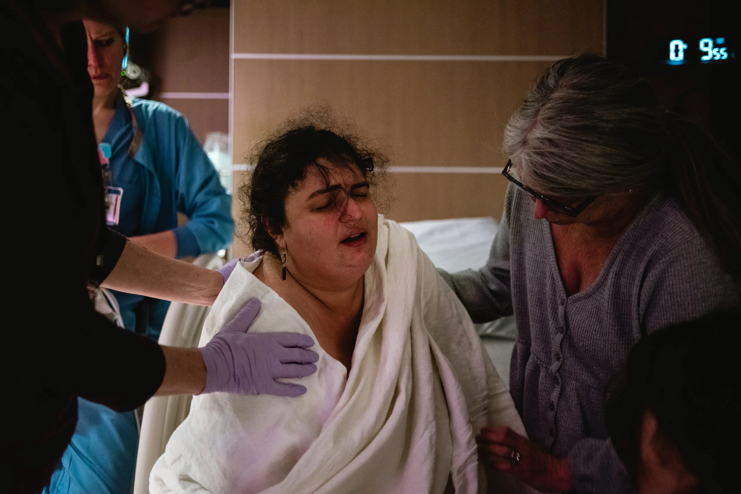  Lucy is at the center of the image, wrapped in a blanket, brow furrowed and mouth open as she moves through a contraction. Curls fall in front of her eyes. Janine’s arms reach in from the left side of the image, wrapping around Lucy’s back and shoul