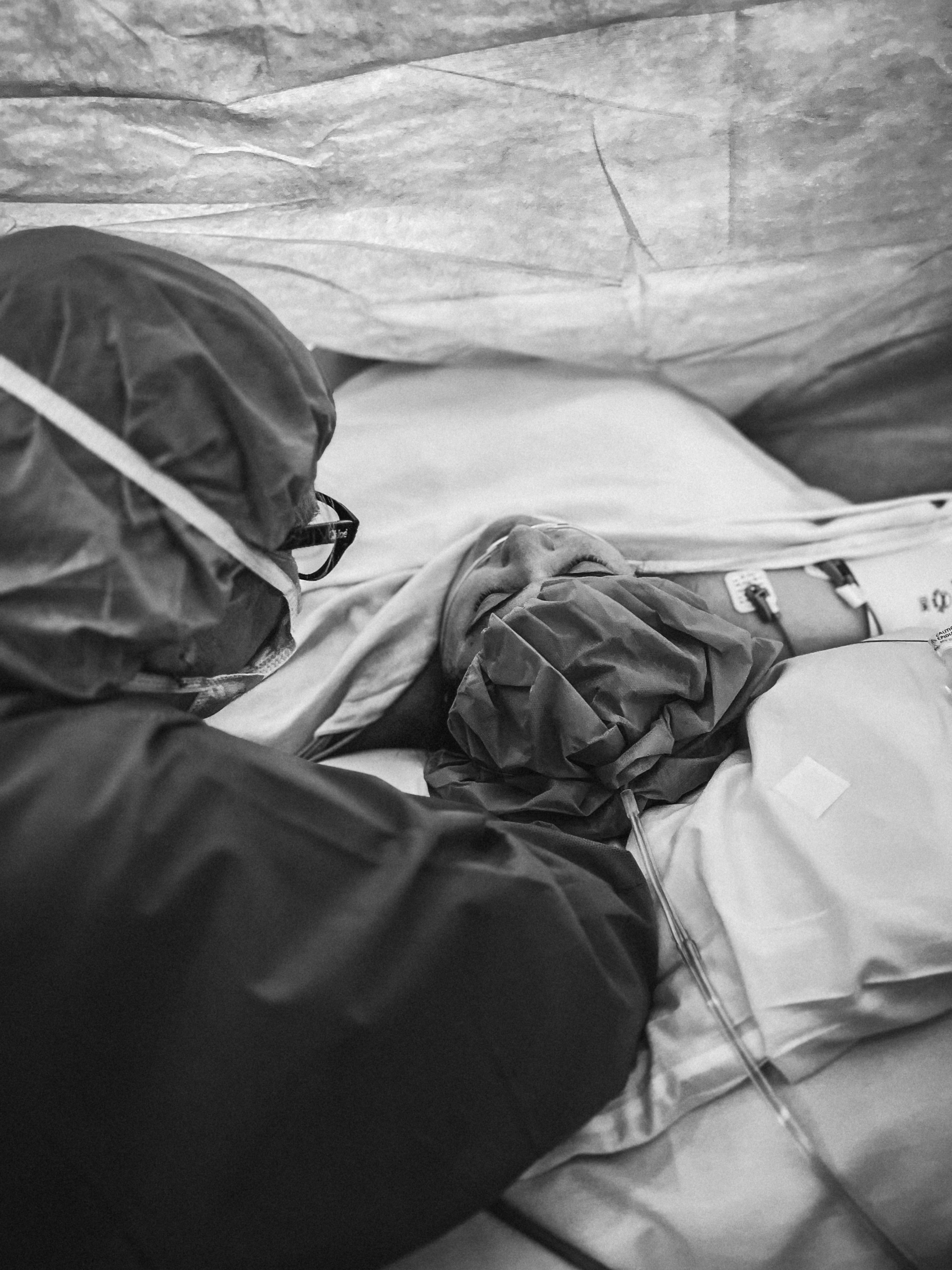  A black and white image of Lucy lying on her back on an operating table. Her chest and head are visible, while the rest of her body is hidden behind a drape. She is wearing a surgical cap on her head, a blanket covering her up to her chin. She has h