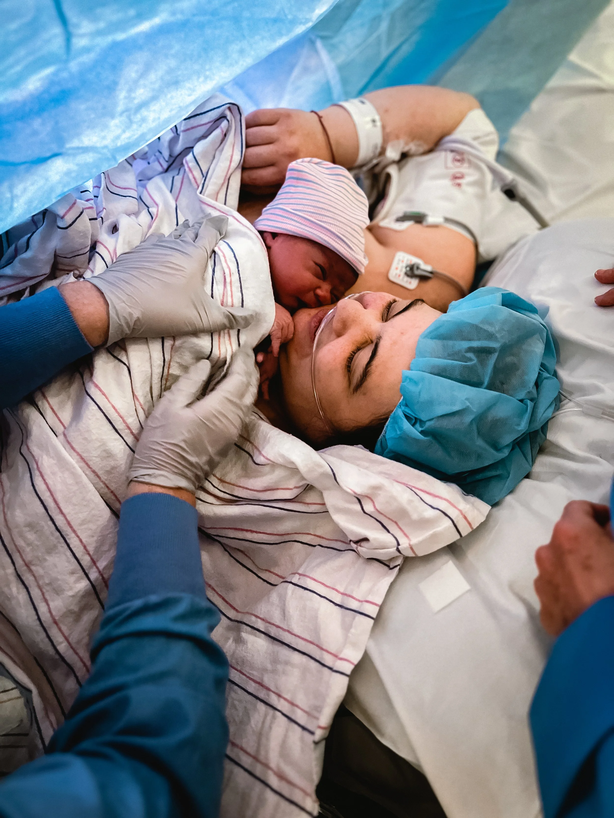  An overhead image of Lucy and Beatrice’s first meeting. A nurse’s gloved hands reach in from the left of the image, placing Beatrice on Lucy’s chest, touching their cheeks together.  Lucy smiles softly. Beatrice wears a pink and blue striped infant 