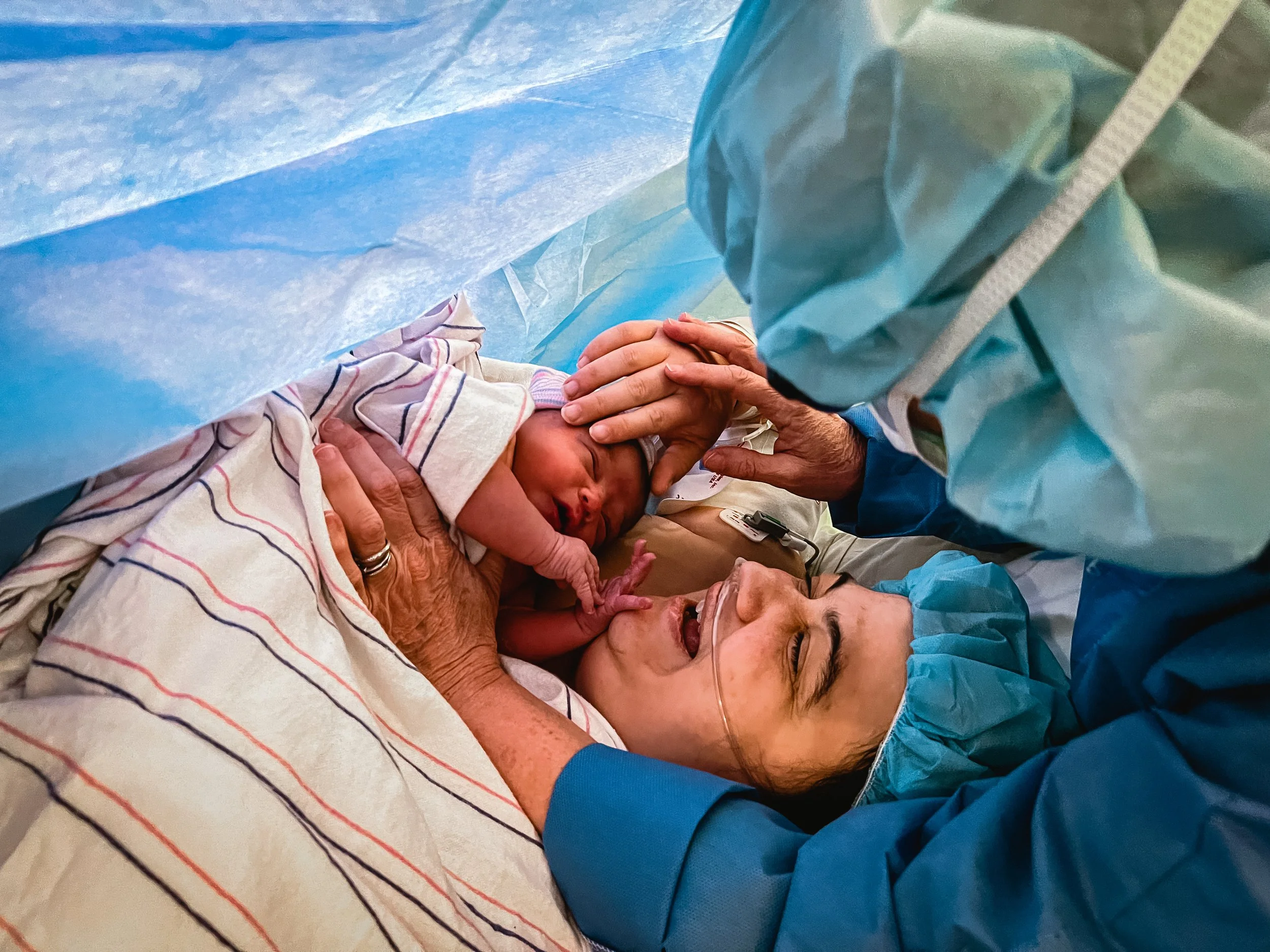  An overhead image of Beatrice and Lucy. Meg reaches in from the right side of the photo, helping to place Lucy’s hand on Beatrice’s face, while using her other hand to hold Beatrice to Lucy’s chest. Lucy’s smile lights up her face.  