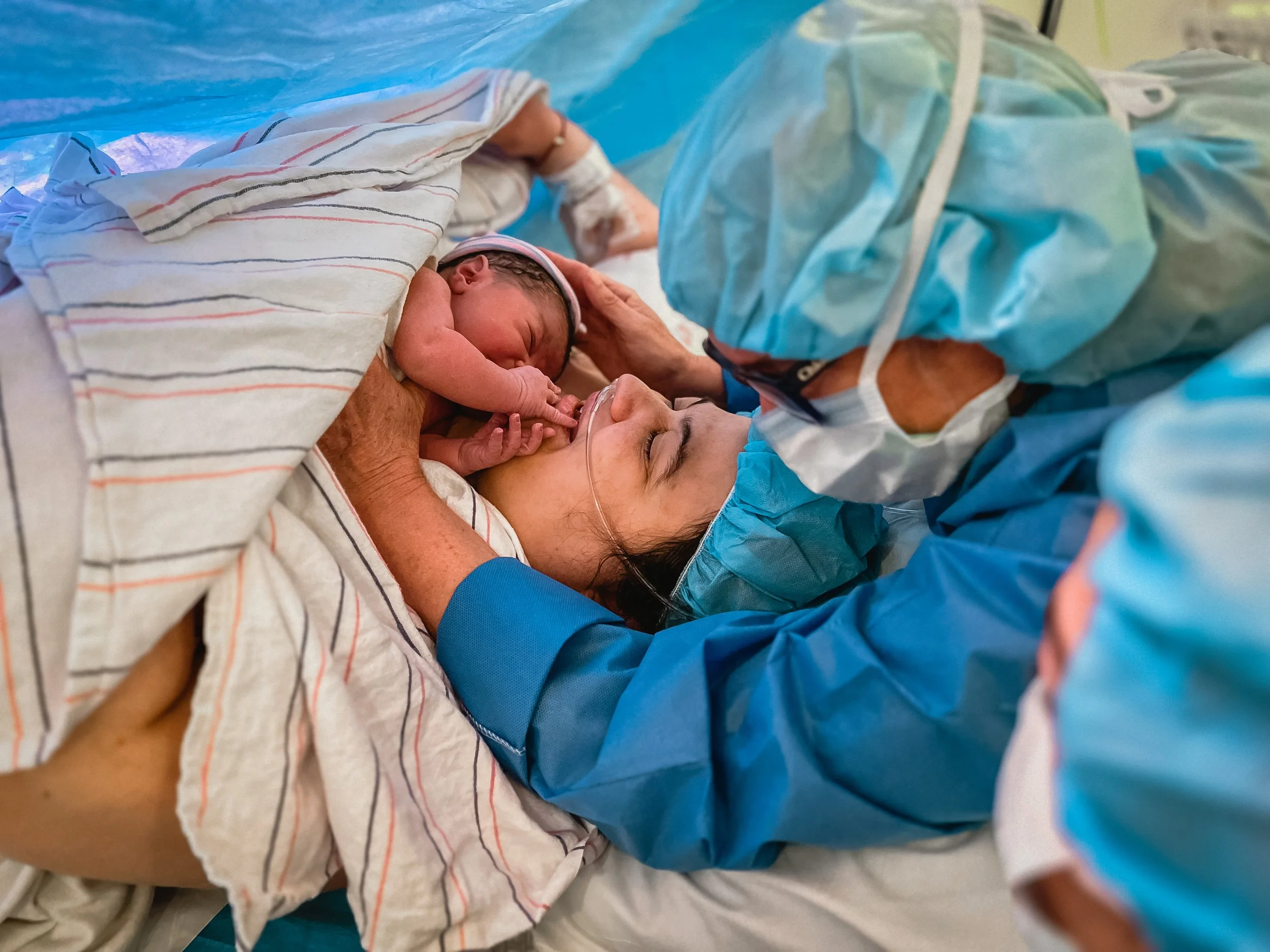  Beatrice reaches with both hands to touch Lucy’s lips, who receives the touch with a smile. Lucy’s arm wraps under a white blanket with blue and pink stripes, holding Beatrice to her chest. Meg leans over Lucy’s head, helping to hold Beatrice to Luc