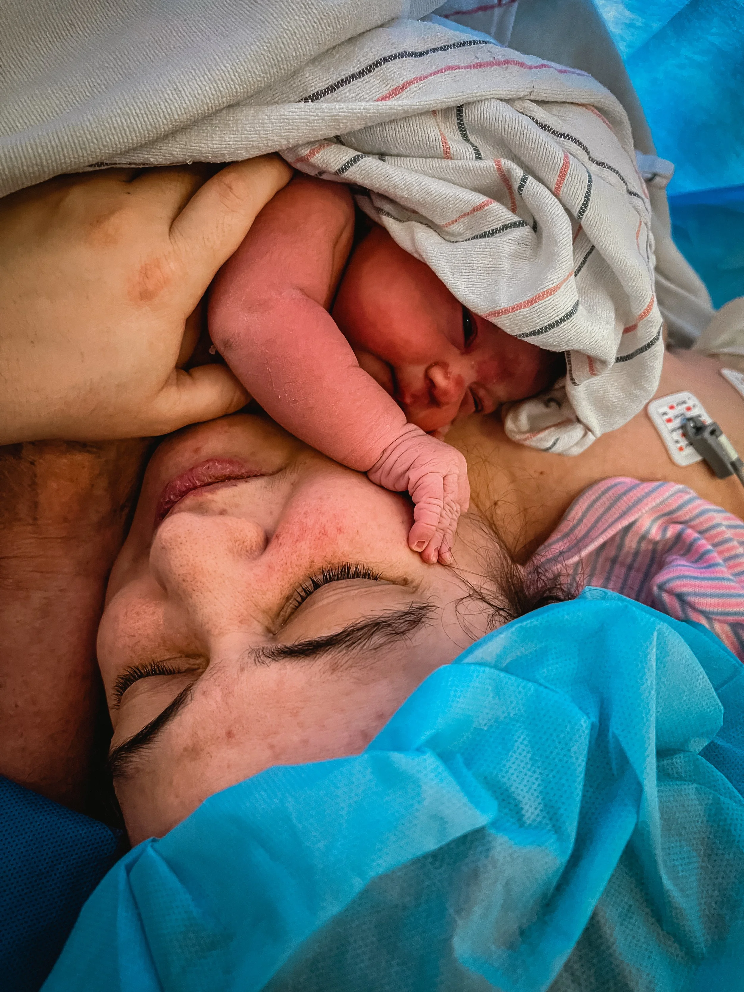  A close-up portrait of Lucy’s face. She is calm, eyes closed, as she tilts her cheek to the left. Her hand is clasped around Beatrice’s chest, whose hand is holding Lucy’s cheek. Beatrice is bundled in newborn blankets, but looks with open eyes towa