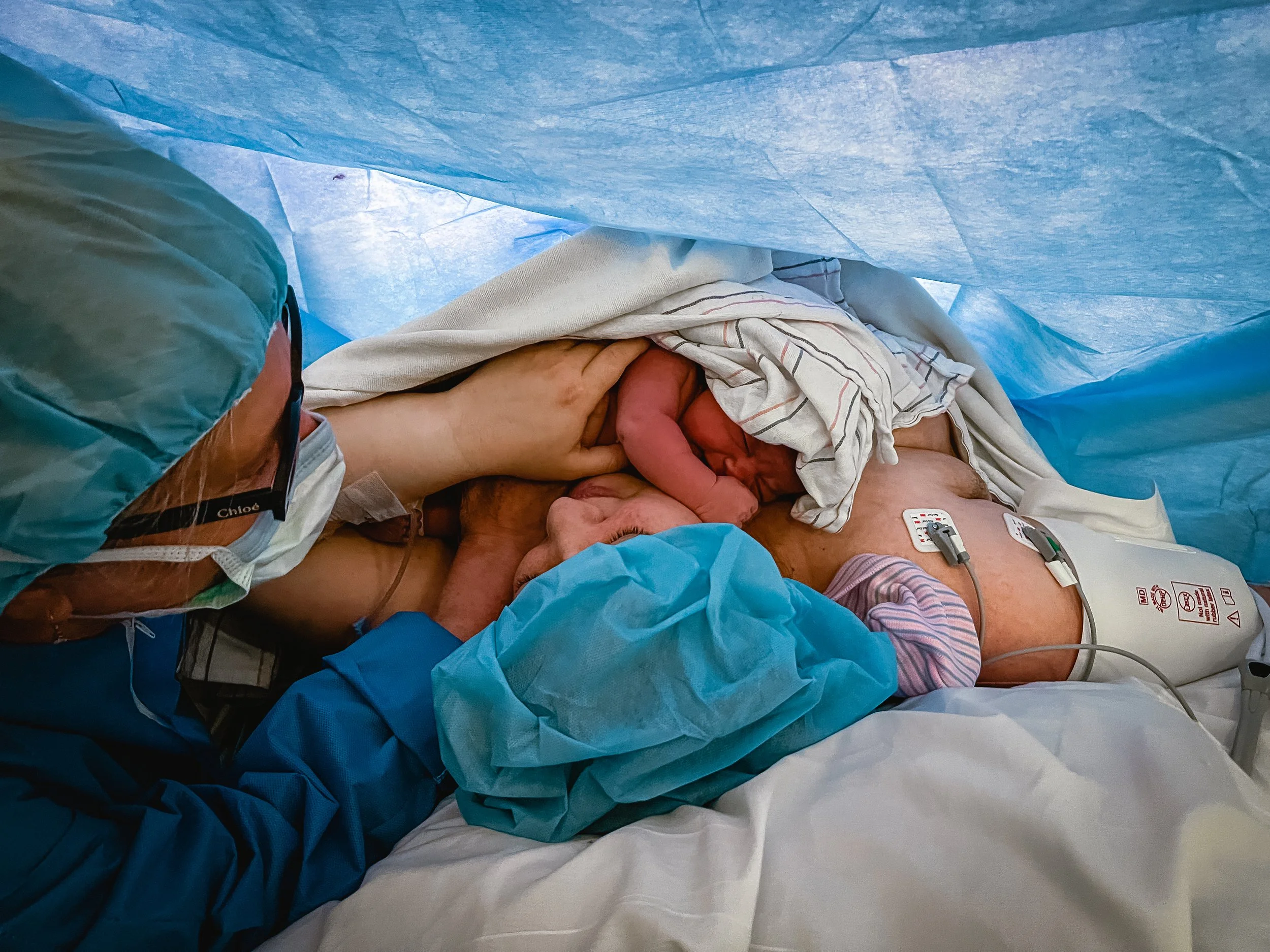  Three generations of women are pictured in an operating room. Meg, a new grandmother, looks at her daughter and granddaughter, a loving hand placed on Lucy’s shoulder. Lucy rests calmly, a hand on Beatrice’s small body, as the youngest of the three 