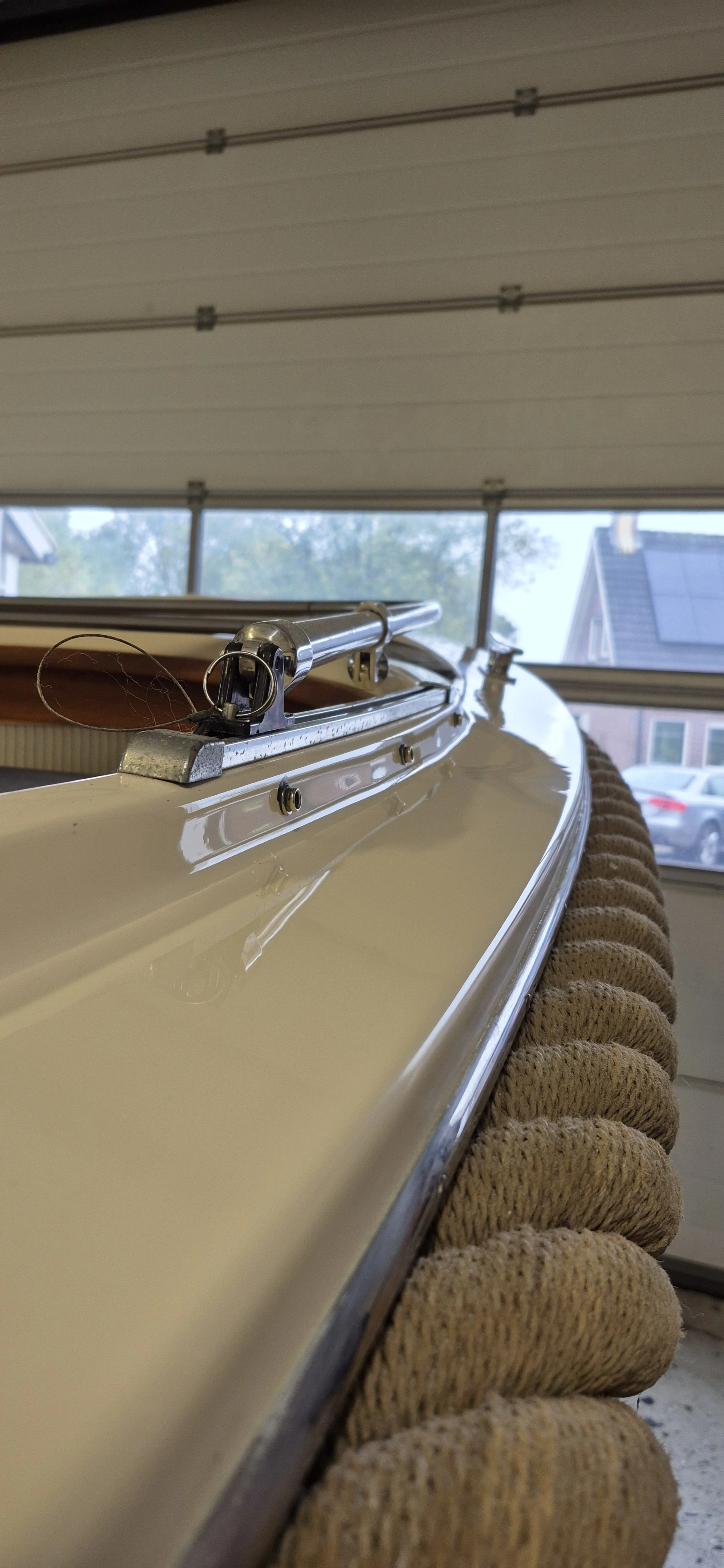 Close-up van een blad met een glanzende witte onderkant, uitgerust met metalen onderdelen. Het is geplaatst op een zachte, gebreide mat in een garage of werkplaats met een raam en auto's buiten.