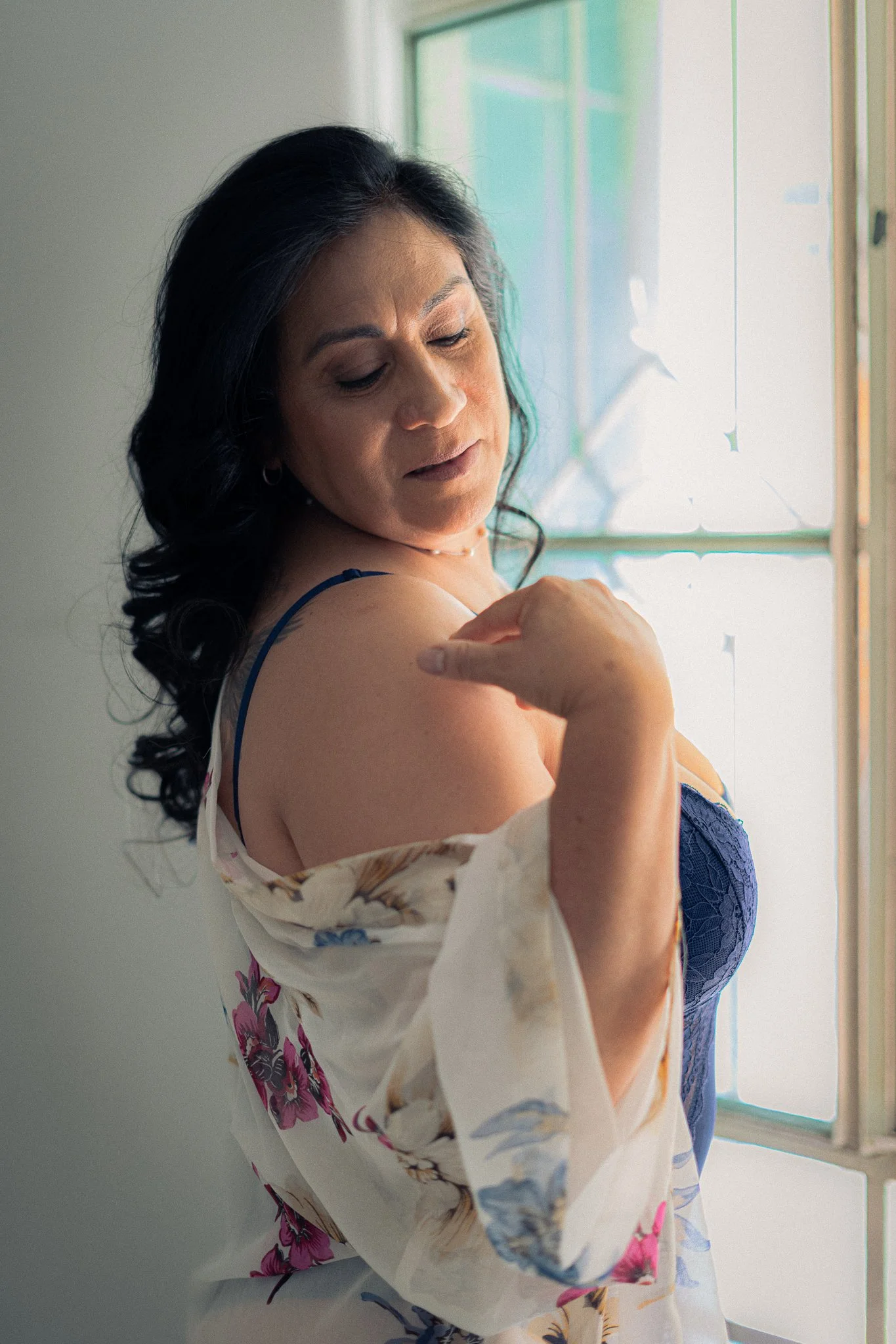 Mujer con cabello oscuro y piel morena mostrando su hombro con una expresión serena, de pie cerca de una ventana con luz natural en un ambiente interior.