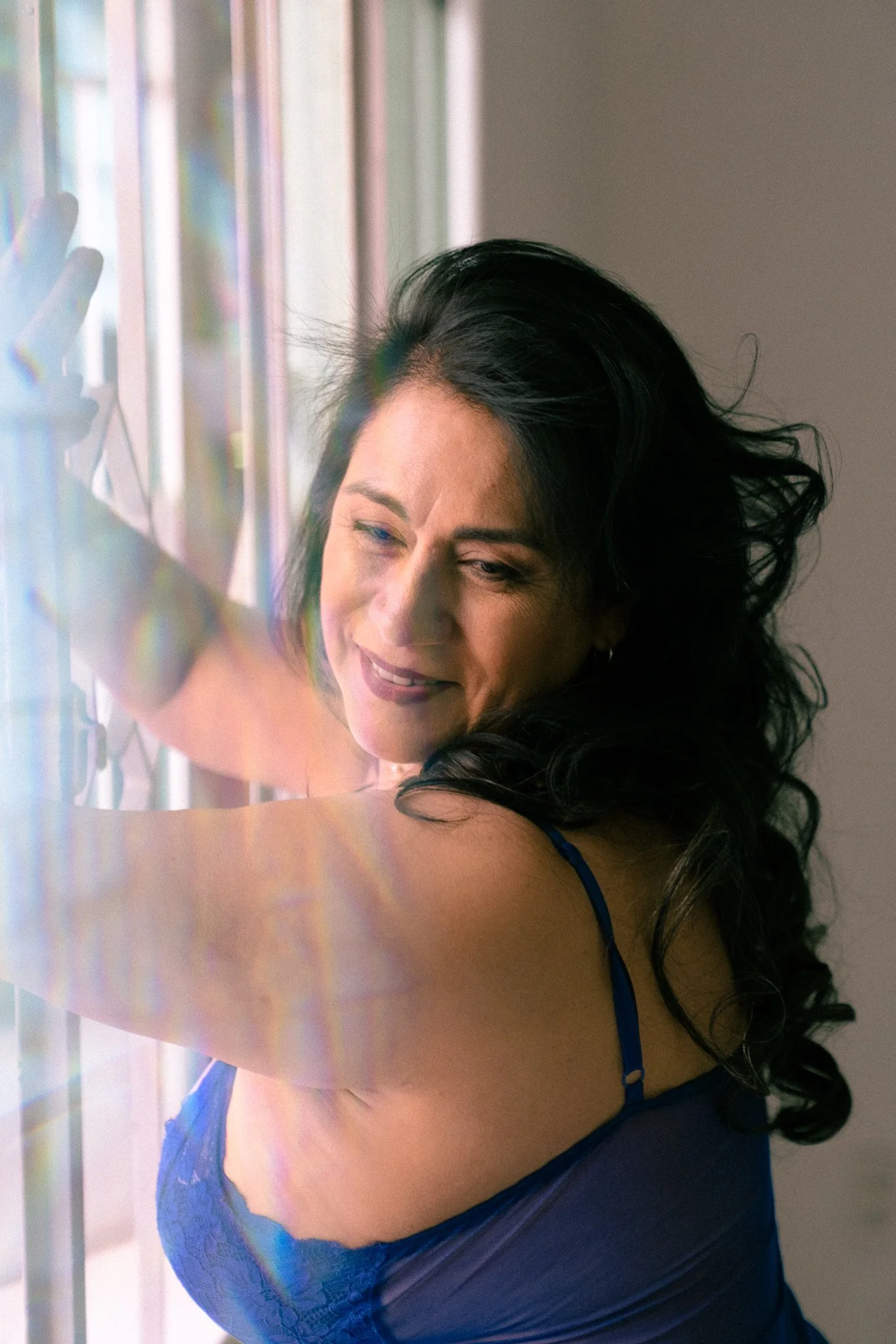 Mujer con cabello rizado negro, vestido azul de encaje, tocando una ventana o espejo con la mano izquierda y sonriendo.