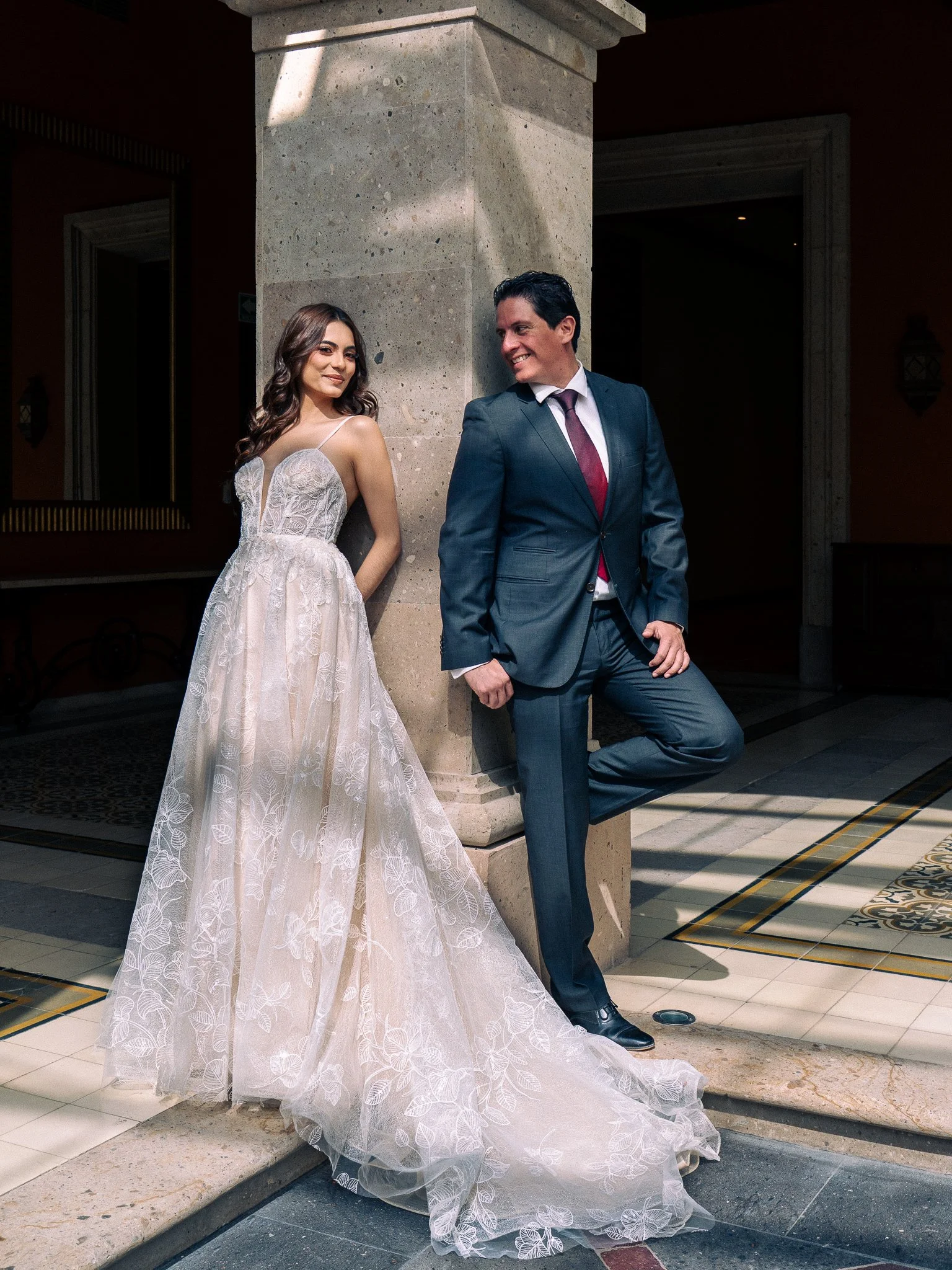 Pareja de novios en un encuadre romántico, la mujer usa un vestido de novia y el hombre traje, posando en un edificio con columnas y suelo decorado.