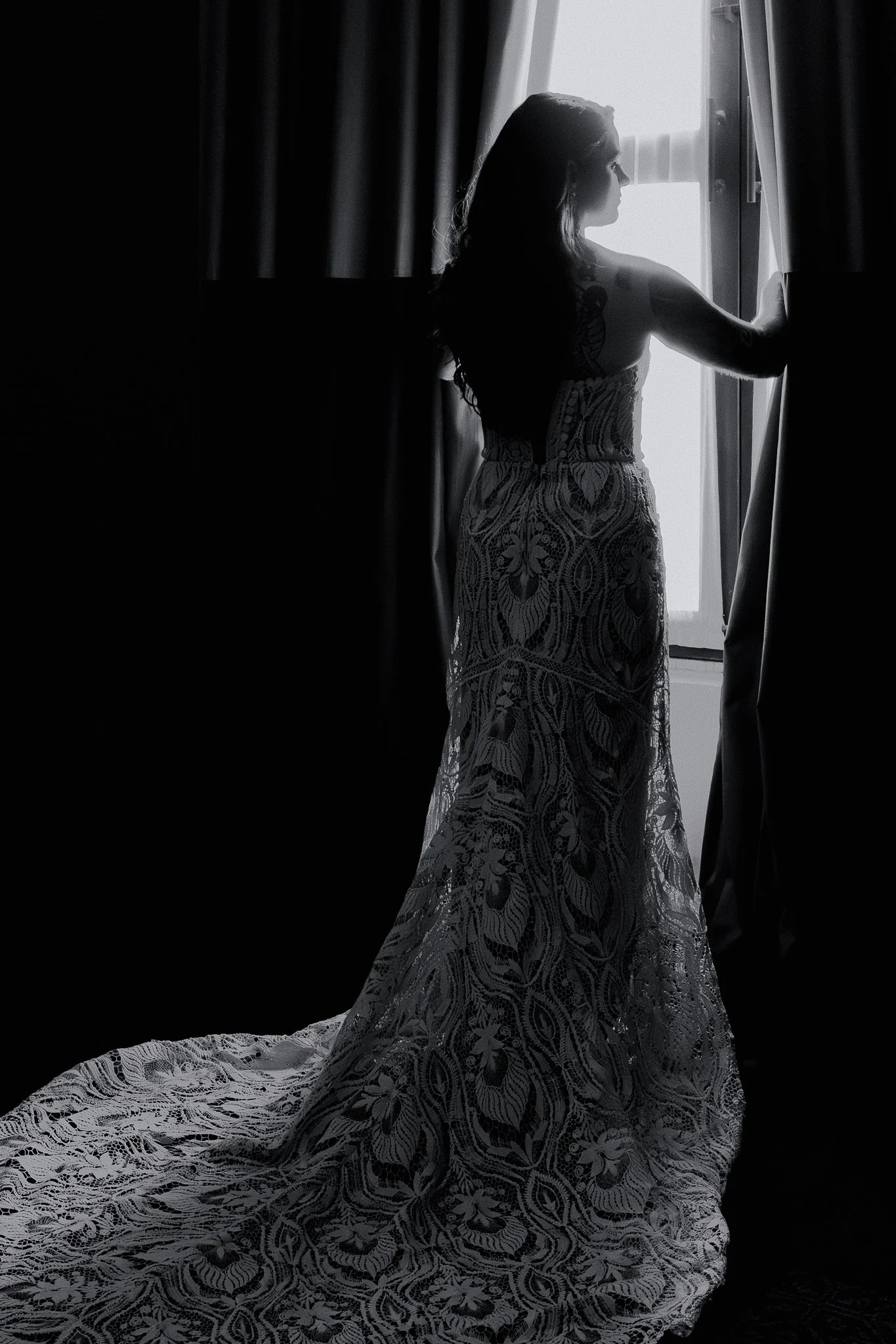 Mujer con vestido de novia en silueta en una habitación, de espaldas, mirando por la ventana mientras sostiene las cortinas, en blanco y negro.