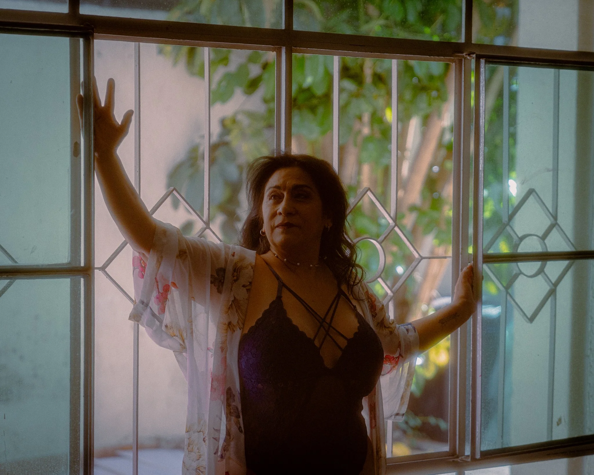 Mujer de cabello castaño oscuro, usando un vestido negro y un kimono blanco con estampado, de pie junto a una ventana, con árboles visibles en el exterior, posando con una mano sobre la ventana.