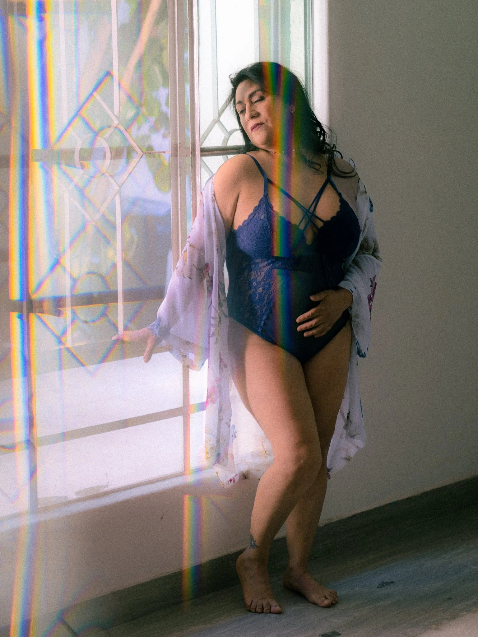 Mujer posando junto a una ventana con reflejos de arcoíris, usando ropa interior y una bata ligera