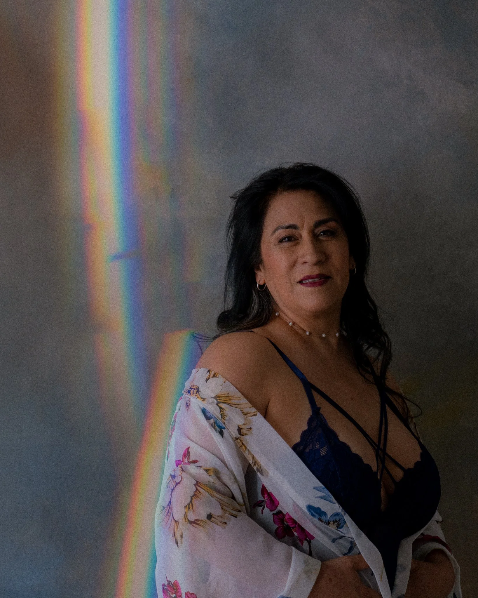 Mujer con blusa de encaje azul y ropa con estampado floral, de cabello oscuro y peinada suelta, posando frente a una pared con efecto de arcoiris de luz